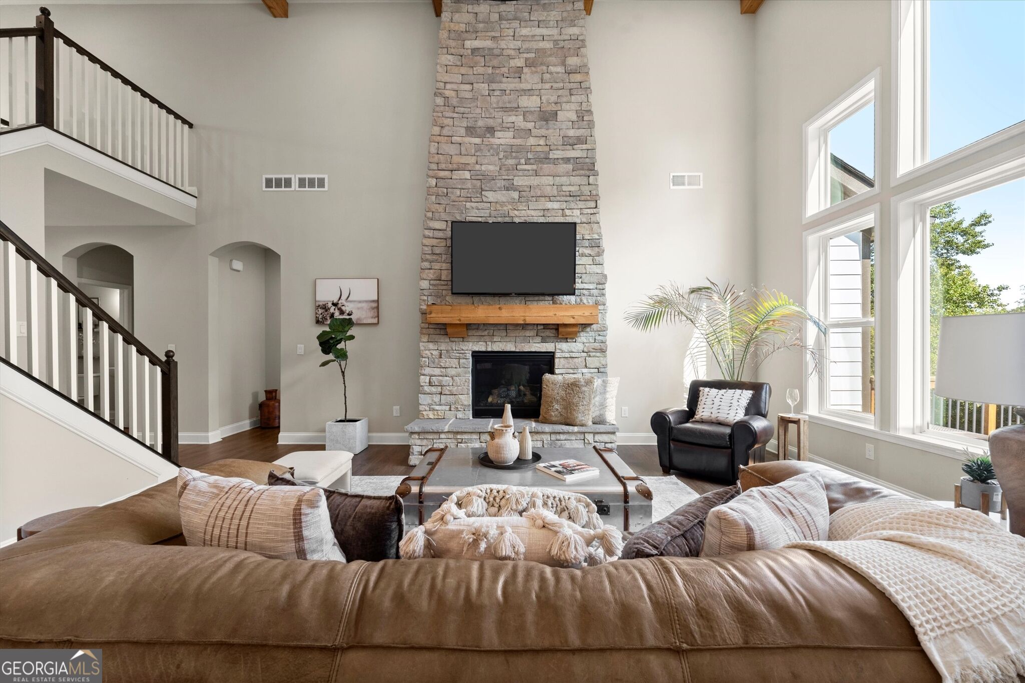 Furnished living room with a tall stone fireplace, sweeping staircase, and large sunlit windows.