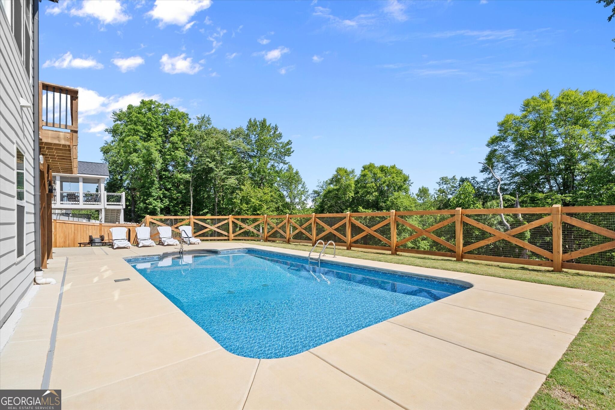Outdoor pool with a wide concrete deck, white lounge chairs, and a wooden fence.