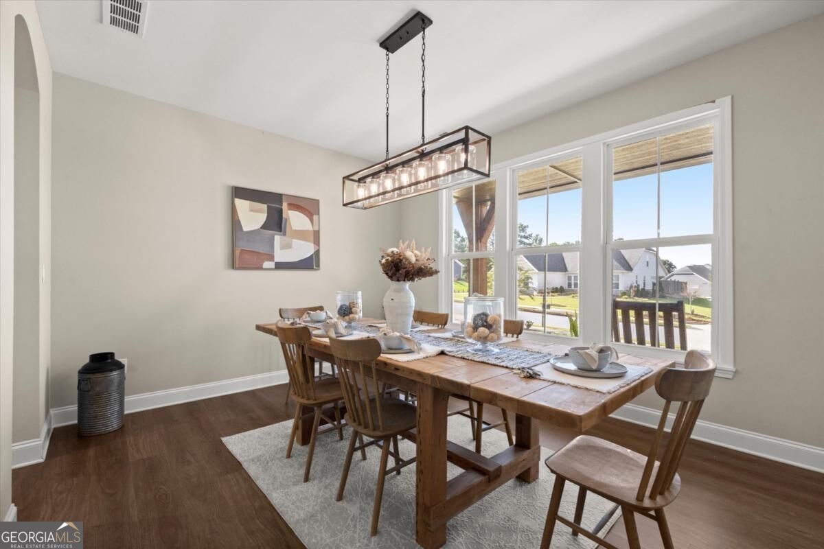 Dining room with a long wooden table, pendant light, and three large windows overlooking neighboring homes.