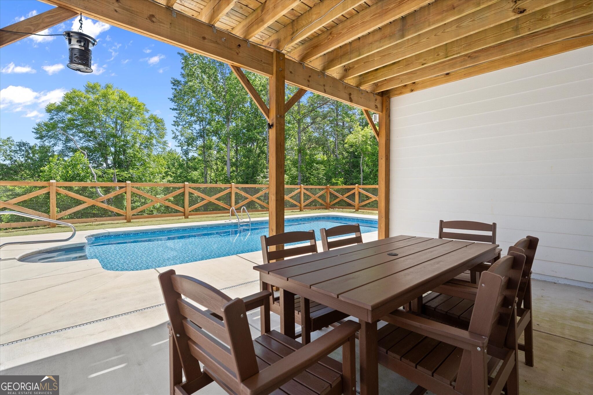 Backyard pool beside a covered timber patio with outdoor dining furniture and a wooded backdrop.