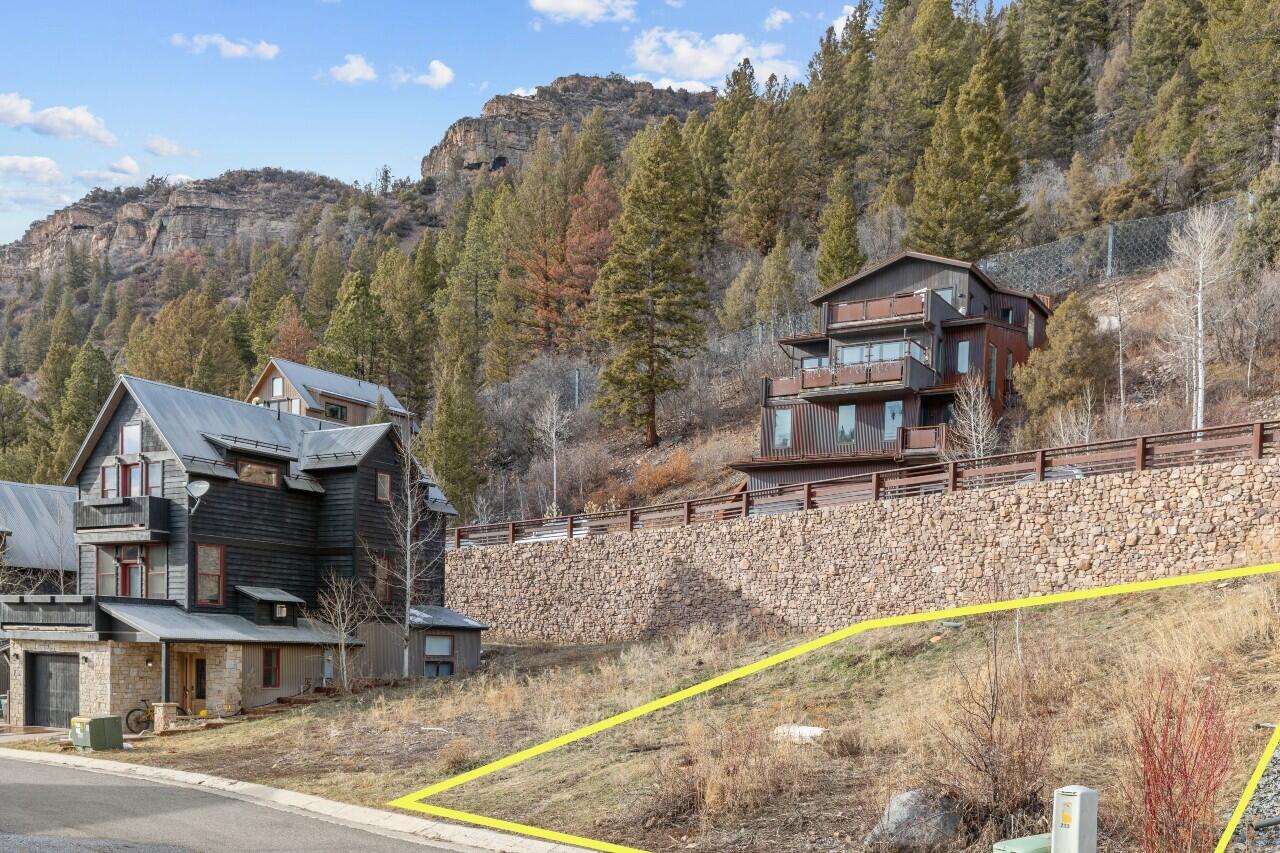 Vacant hillside lot outlined in yellow below mountain homes, beside a stone retaining wall.