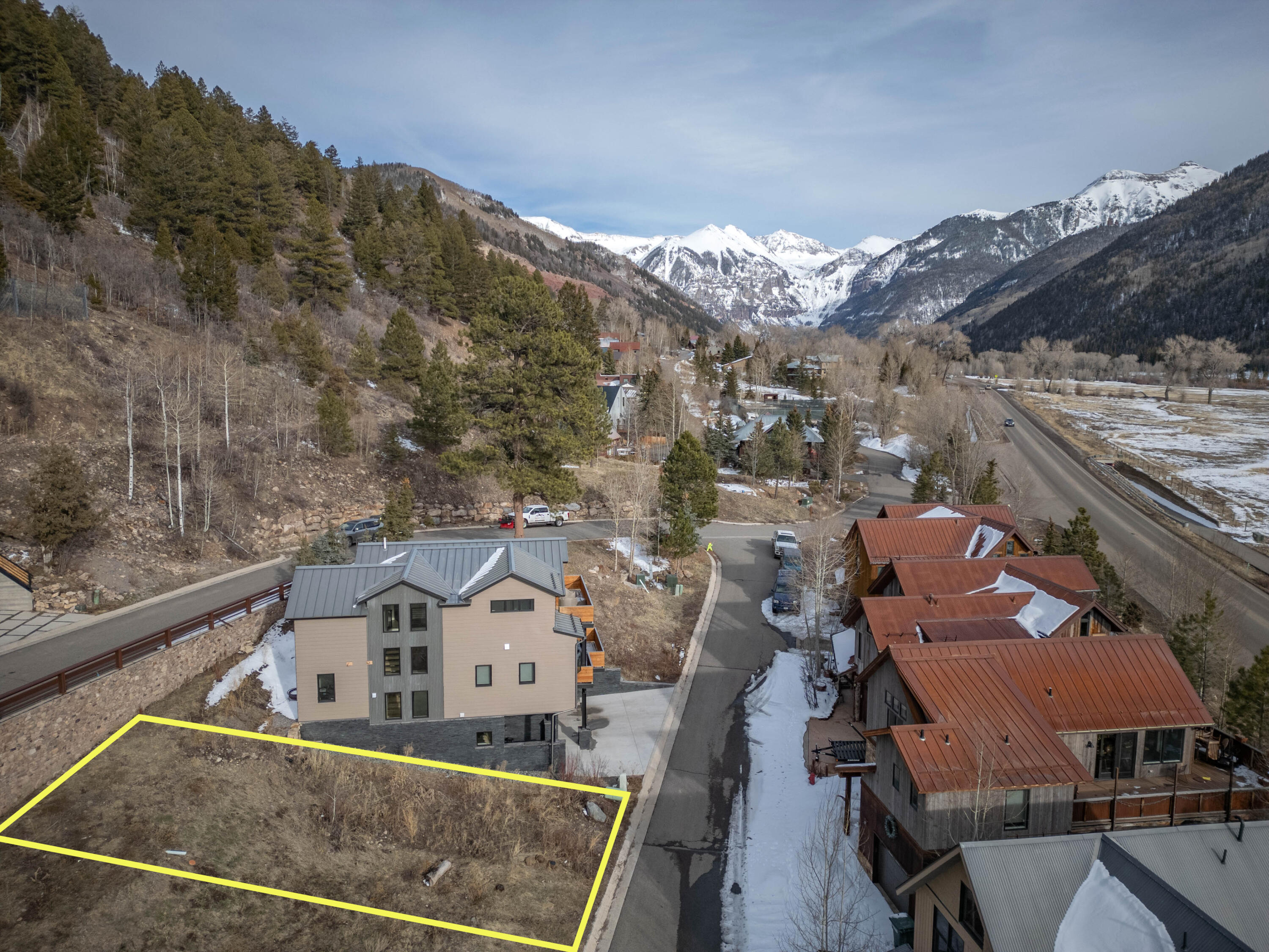 Vacant lot outlined in yellow beside a road, with homes and snow-covered mountains beyond.
