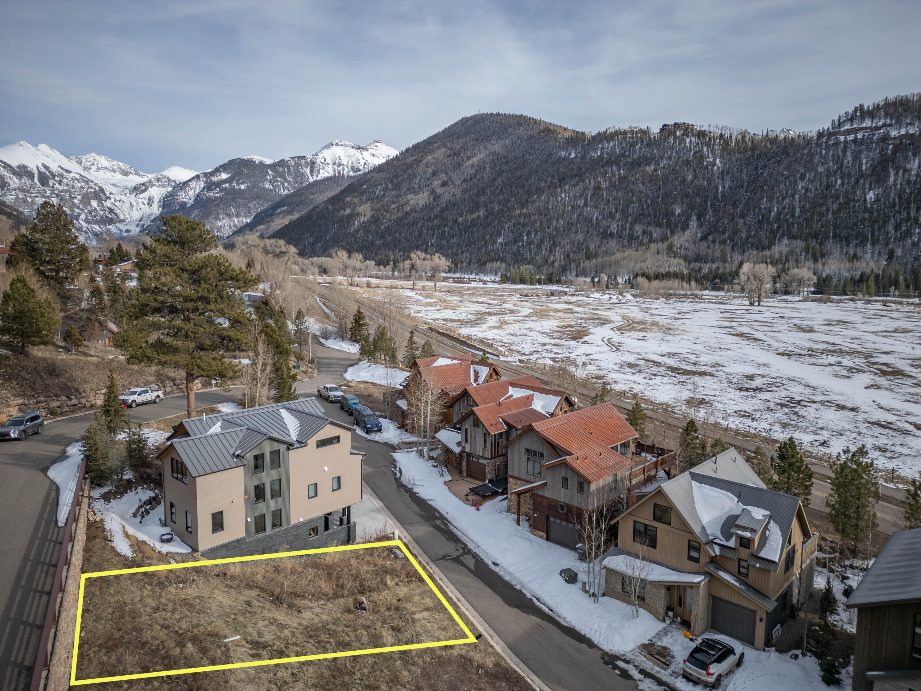 Vacant lot outlined in yellow beside homes, with a snowy valley and mountain views.