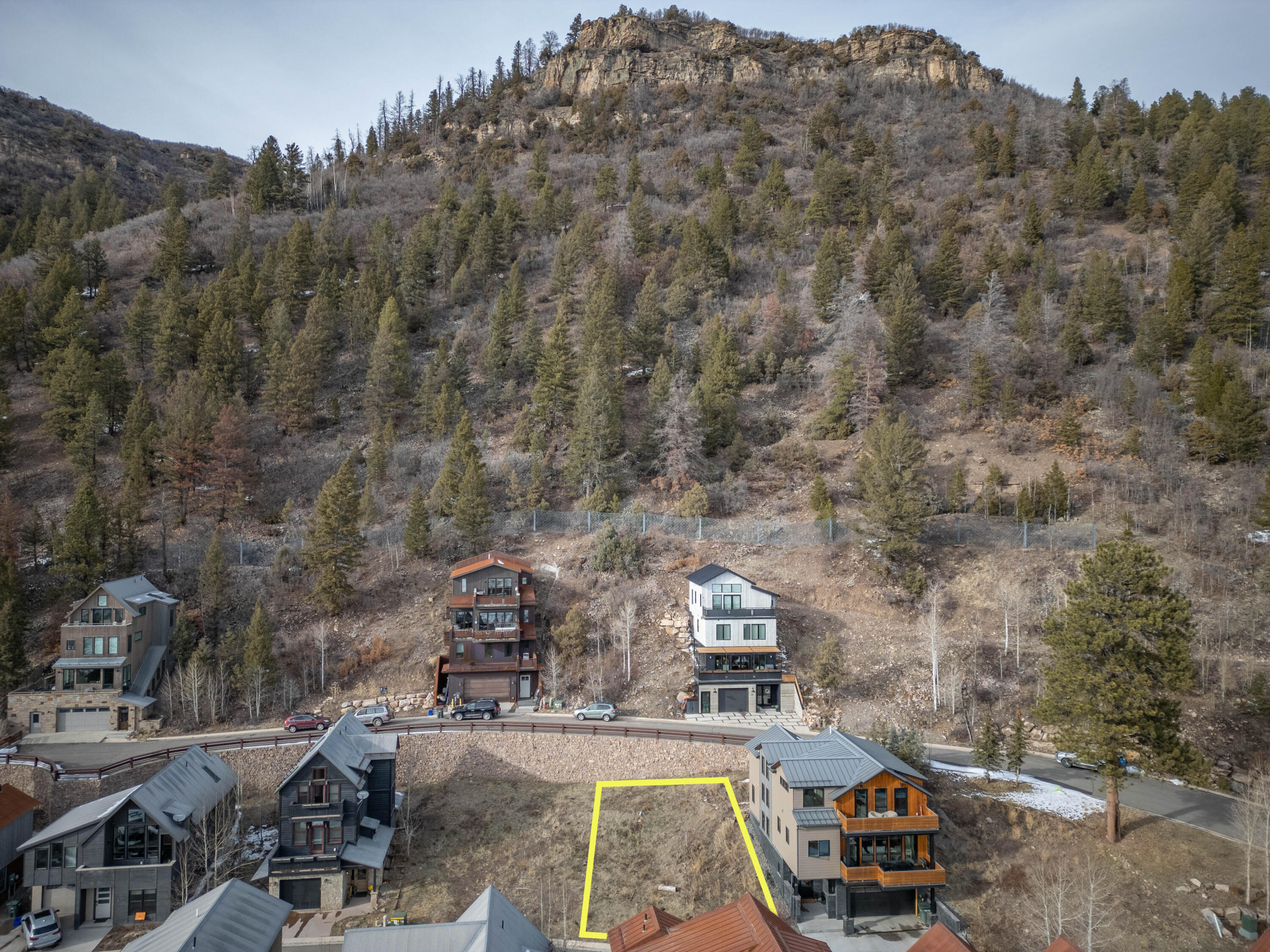 Aerial view of a vacant hillside lot outlined in yellow below a forested mountain.