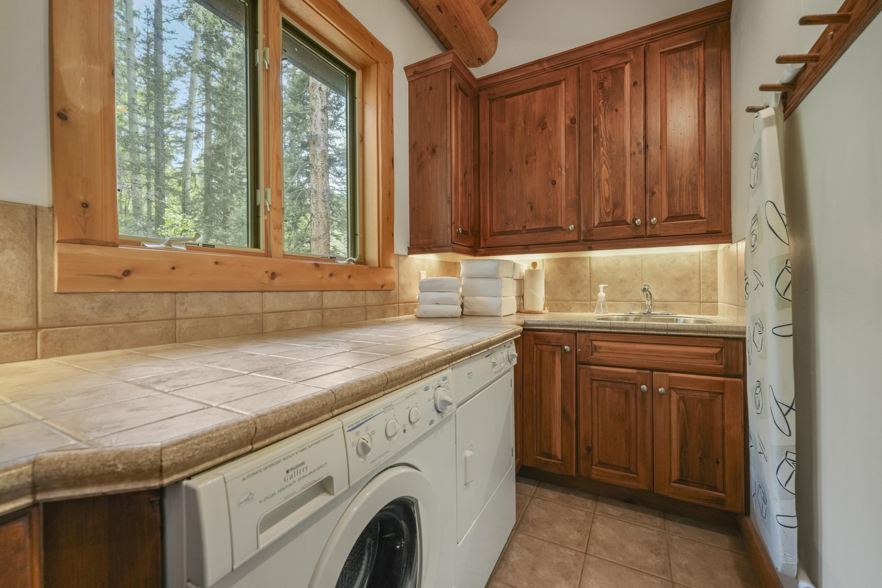 Laundry room with front-loading appliances, tiled counter, wood cabinets, and a window overlooking trees.