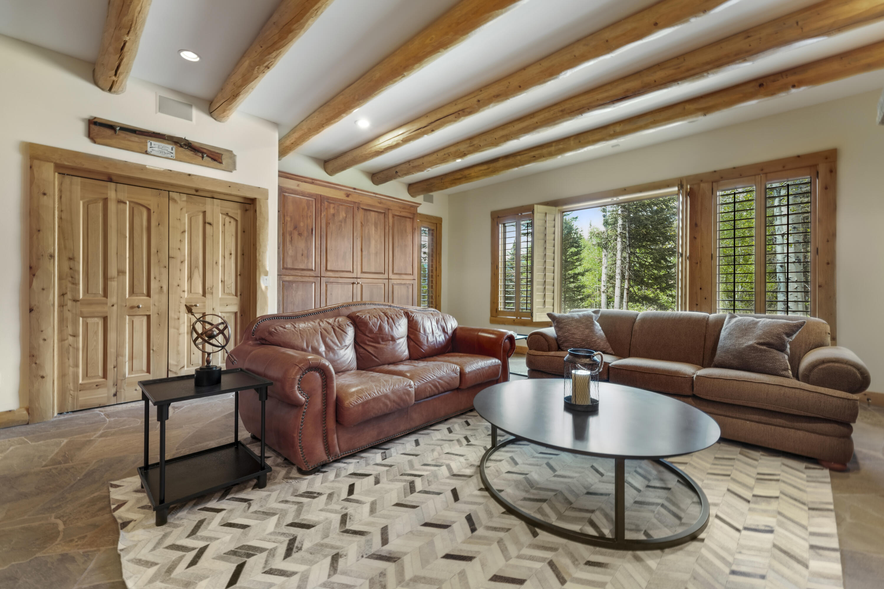 Living room with exposed timber beams, two sofas, and large windows overlooking trees.