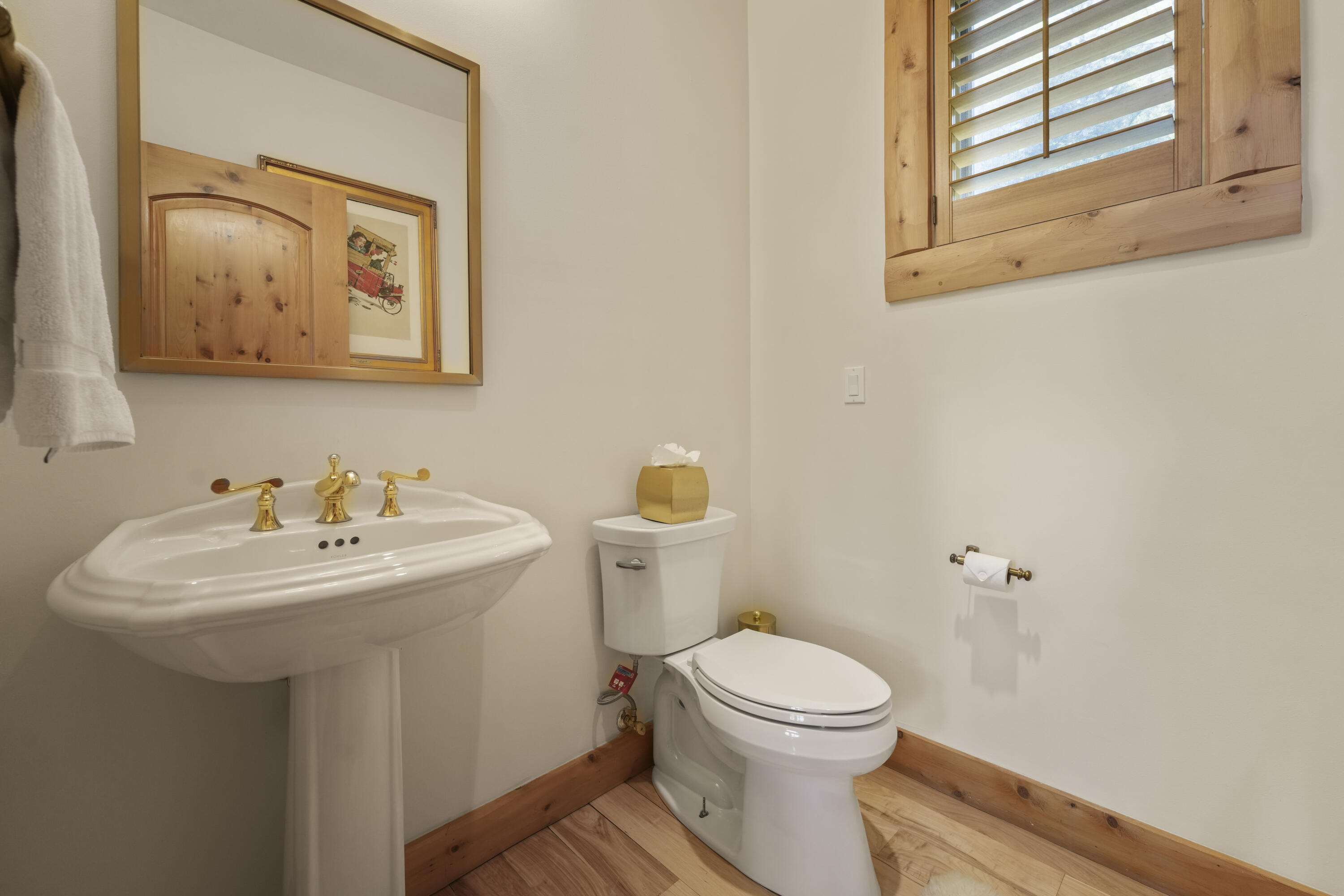 Bathroom with a pedestal sink, toilet, brass fixtures, and a shuttered wood-framed window.