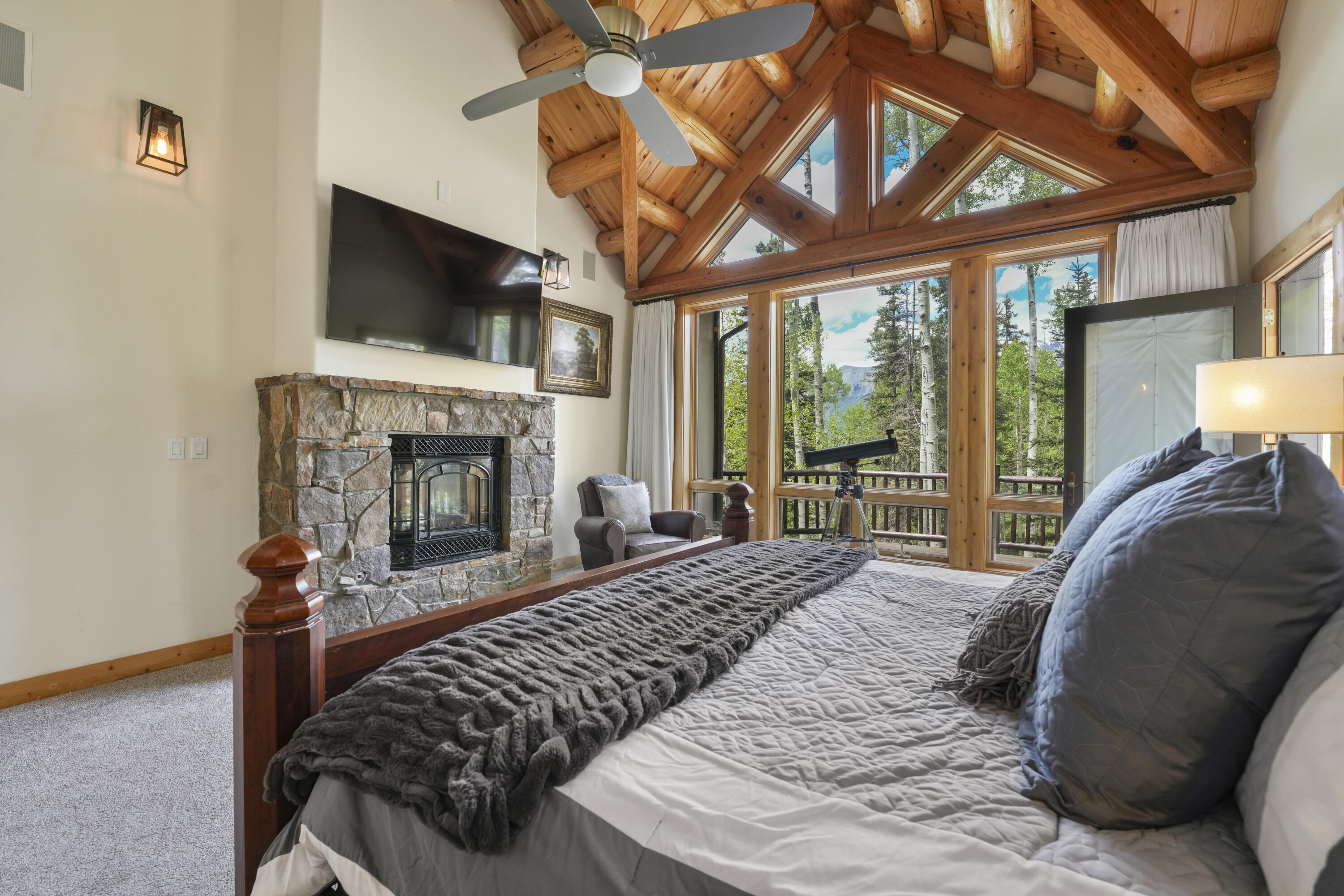 Bedroom with a stone fireplace, vaulted timber ceiling, and large windows overlooking trees and mountains.