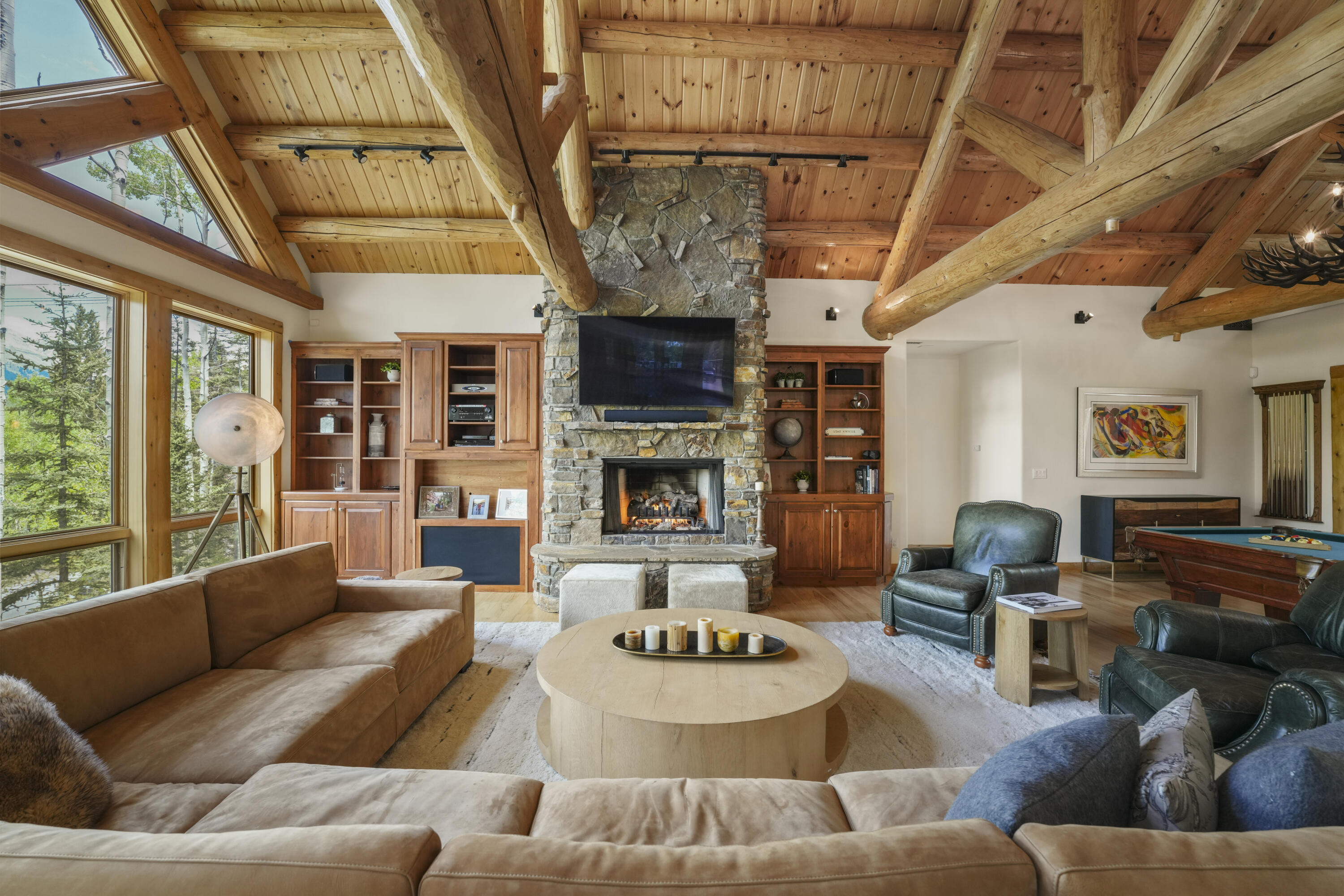 Living room with vaulted timber beams, a stone fireplace, large windows, and an adjoining pool table.