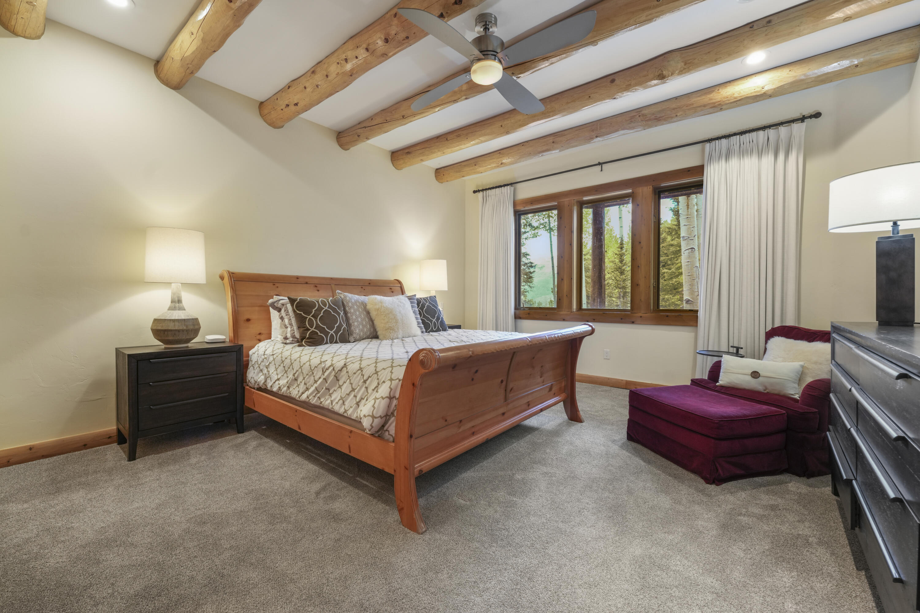 Bedroom with exposed log beams, a wooden bed, and windows overlooking trees.
