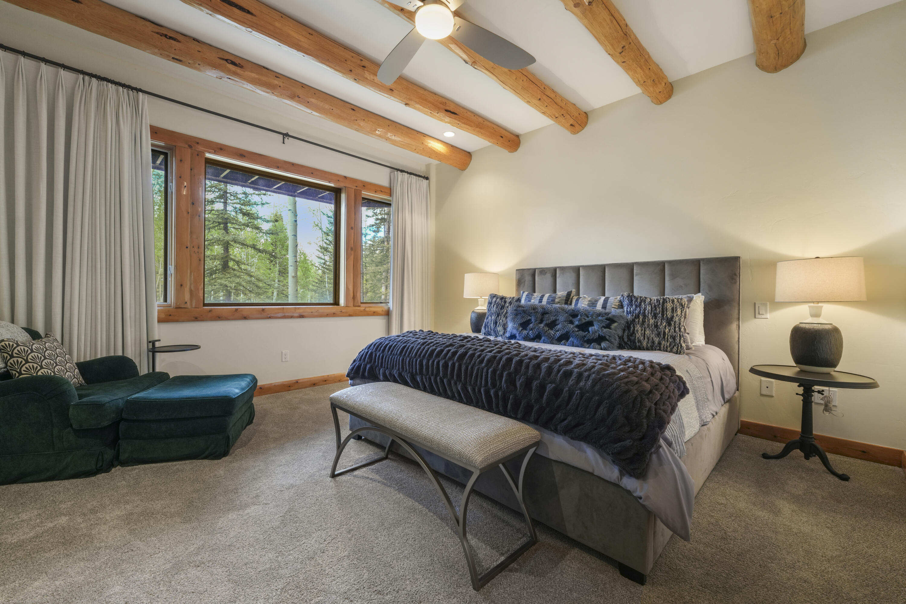 Bedroom with exposed wood beams, a large bed, and windows overlooking trees.
