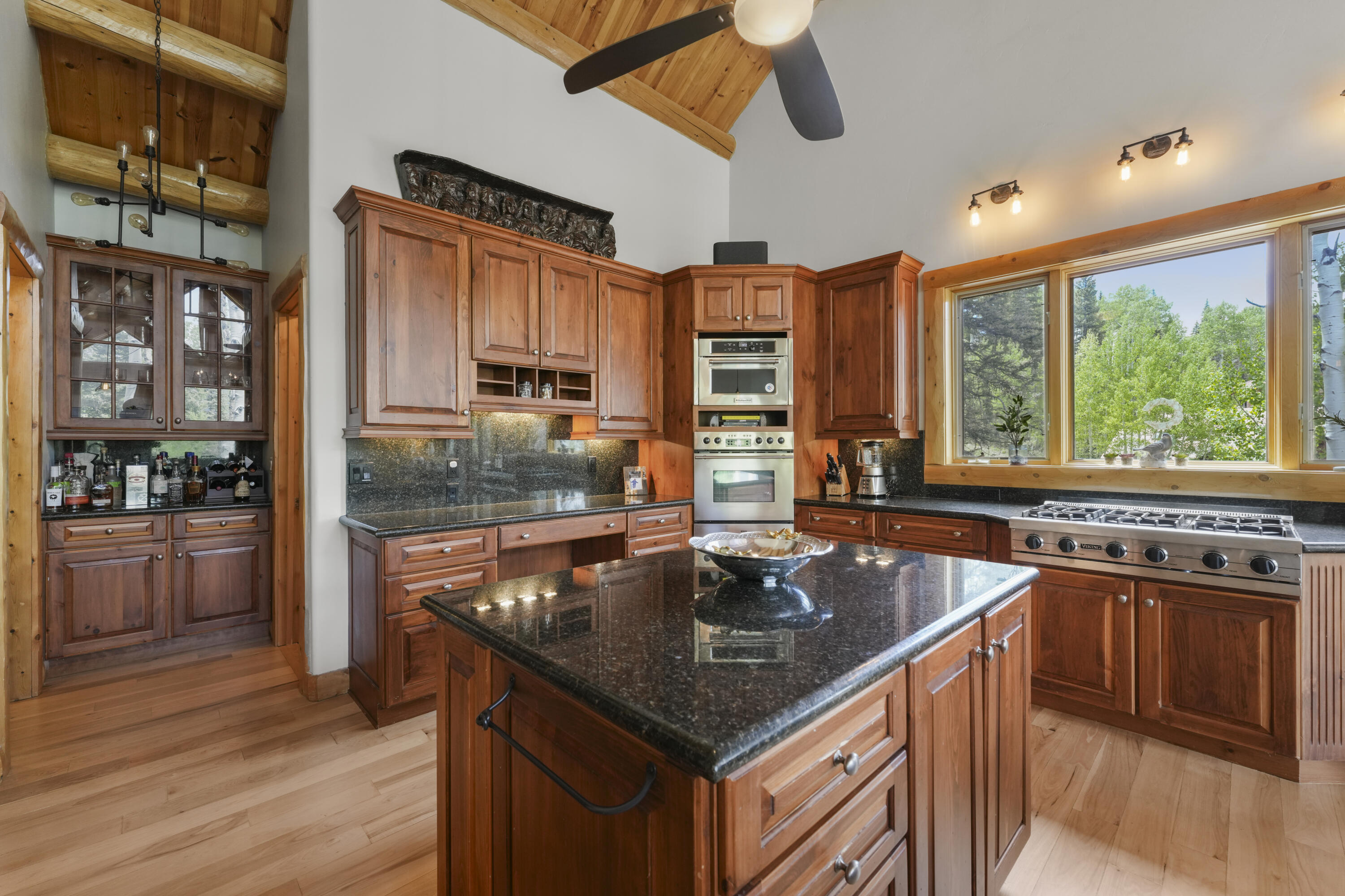 Kitchen with reddish wood cabinets, a granite island, stainless steel appliances, and a broad window overlooking trees.