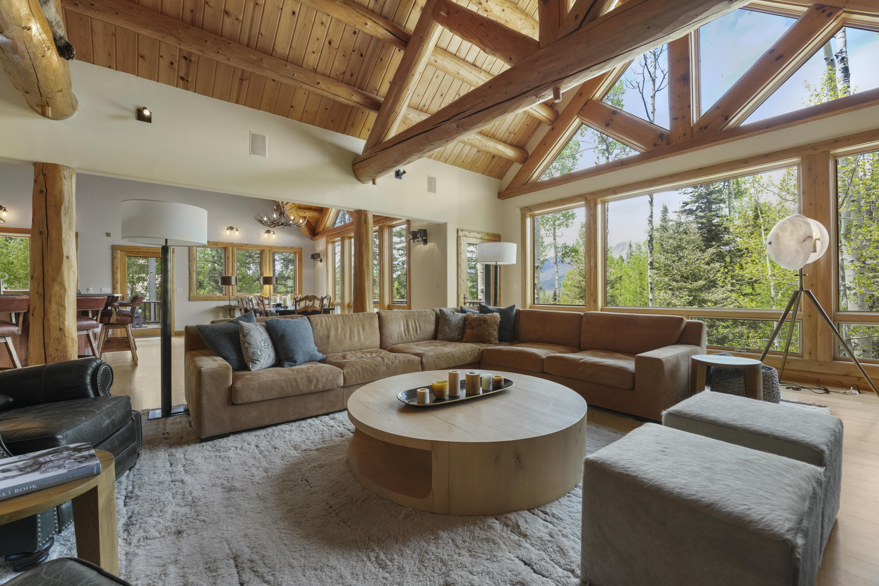 Vaulted living room with exposed timber beams, a sectional sofa, and large windows overlooking trees and mountains.