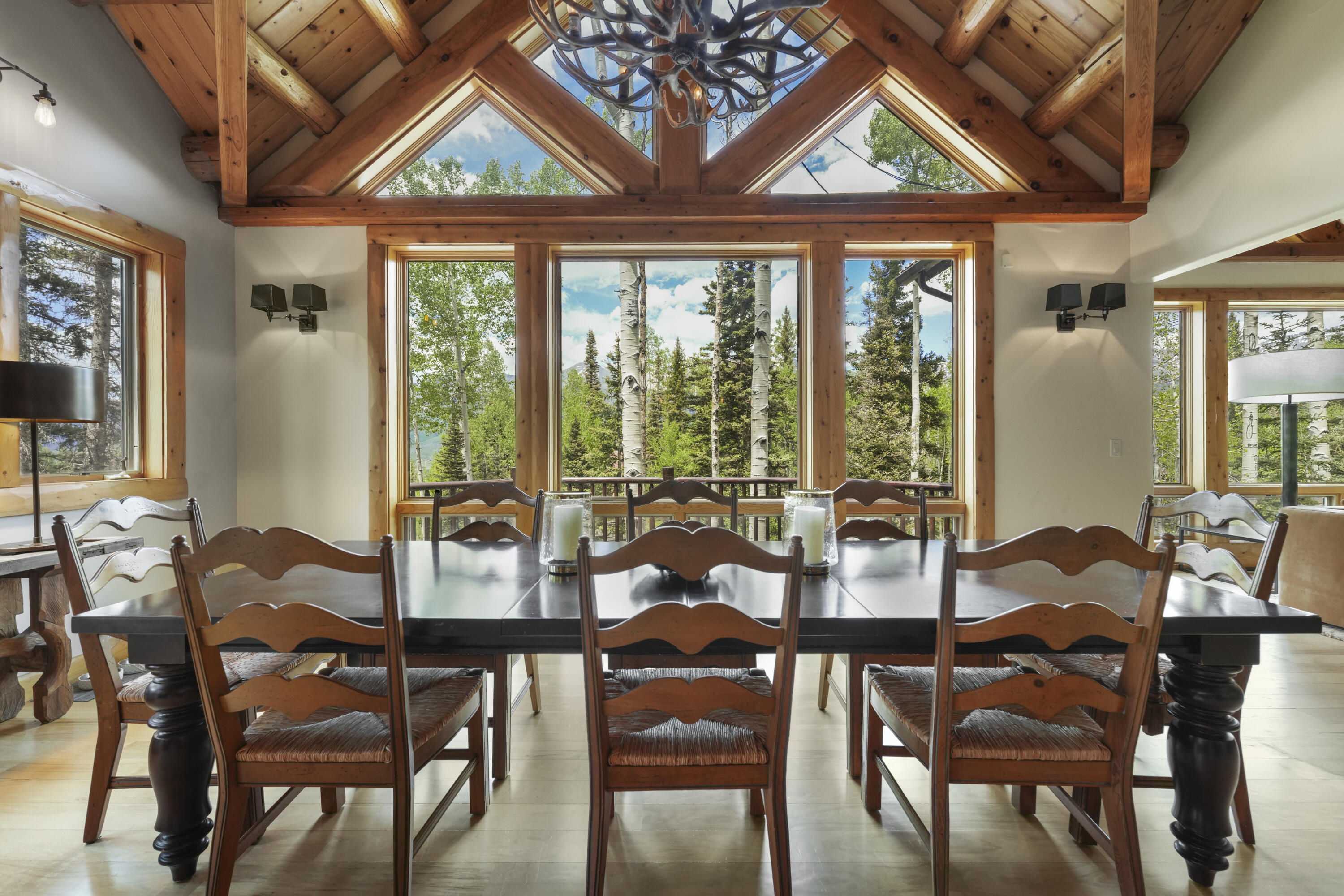 Dining room with a timber vaulted ceiling, antler chandelier, and forest views through large windows.