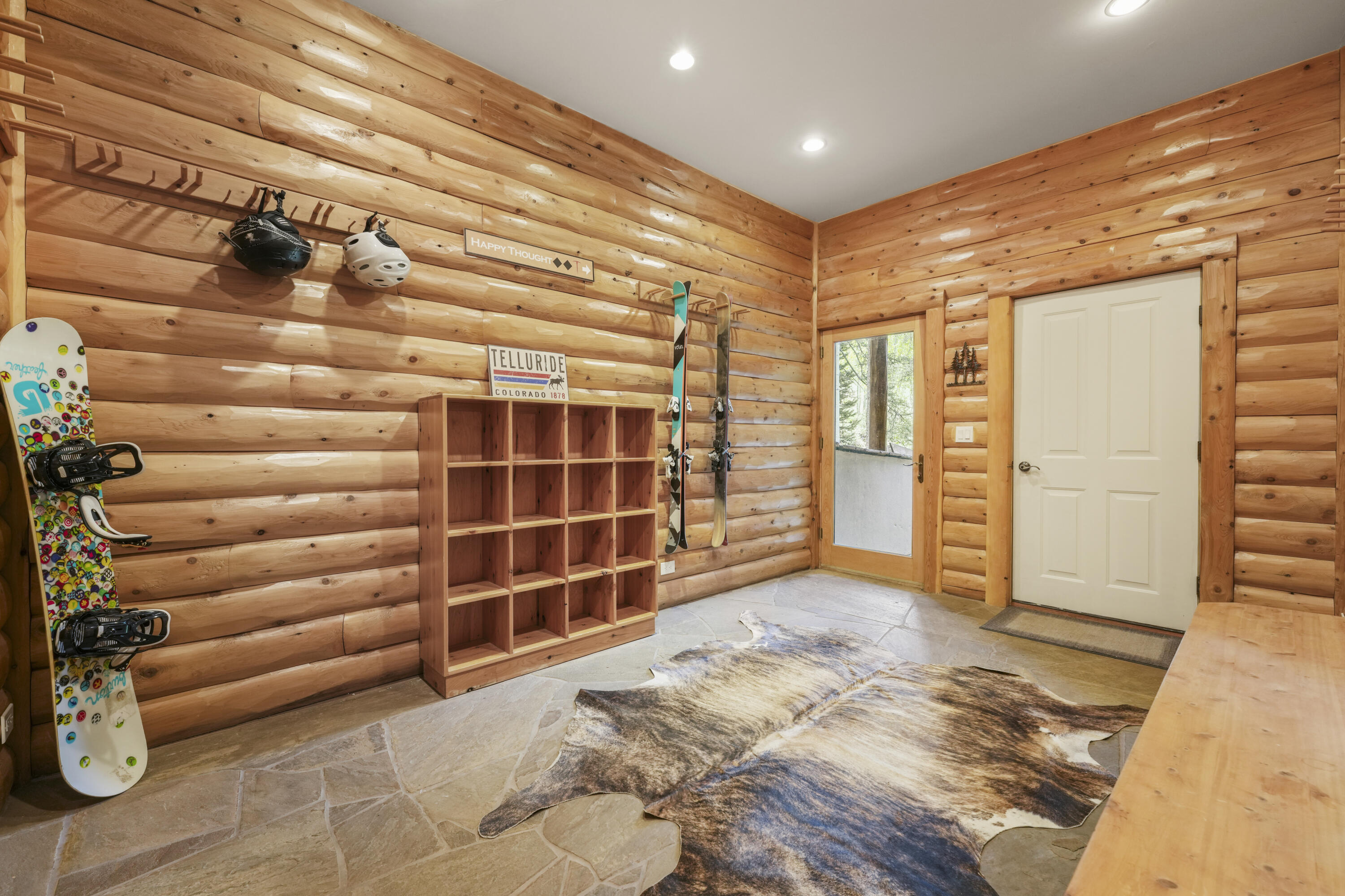 Log-walled ski room with mounted snowboards, helmets, wooden cubbies, and a cowhide rug.