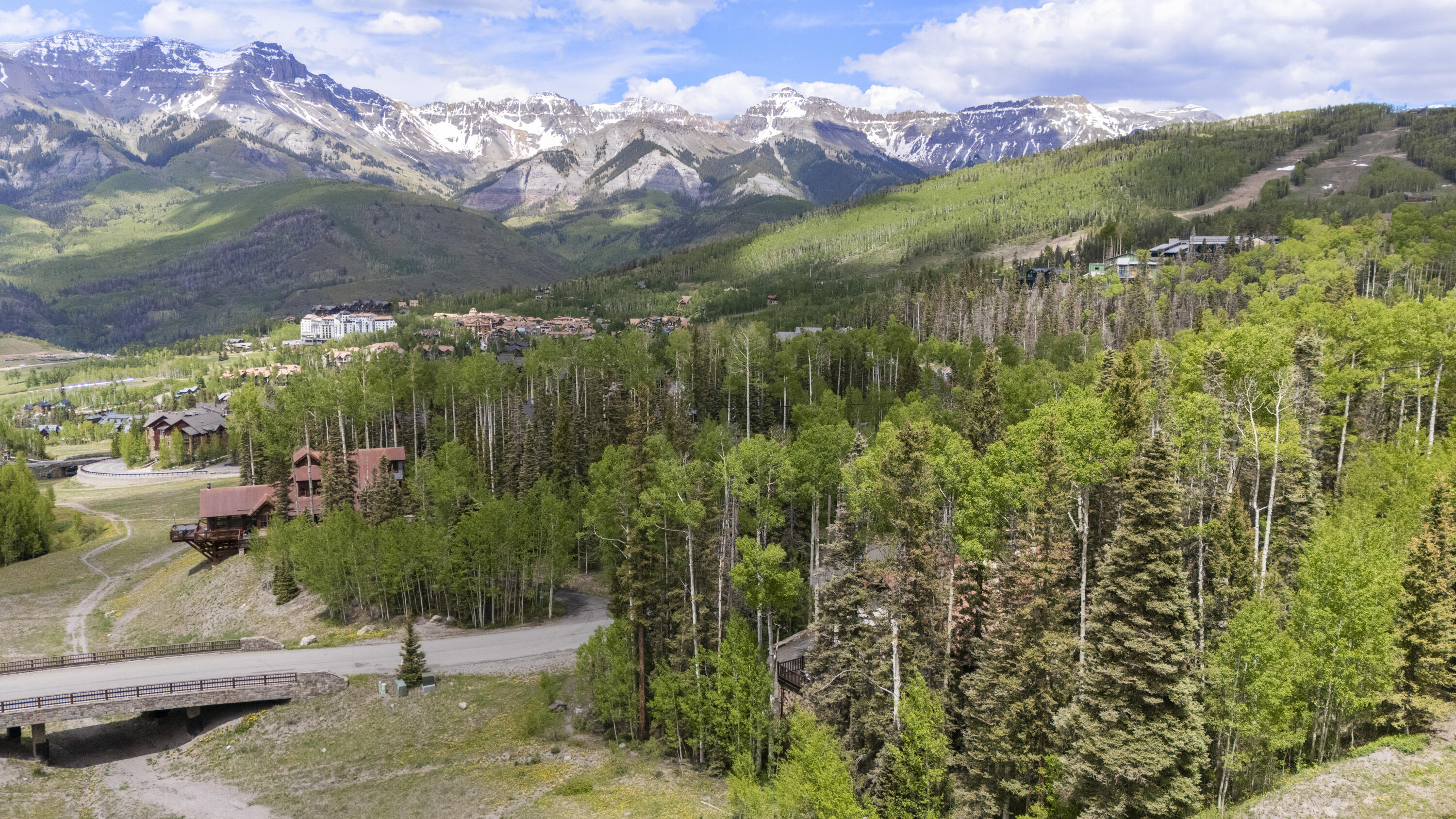 Aerial view of mountain village homes among forest, with snow-streaked peaks in the distance.