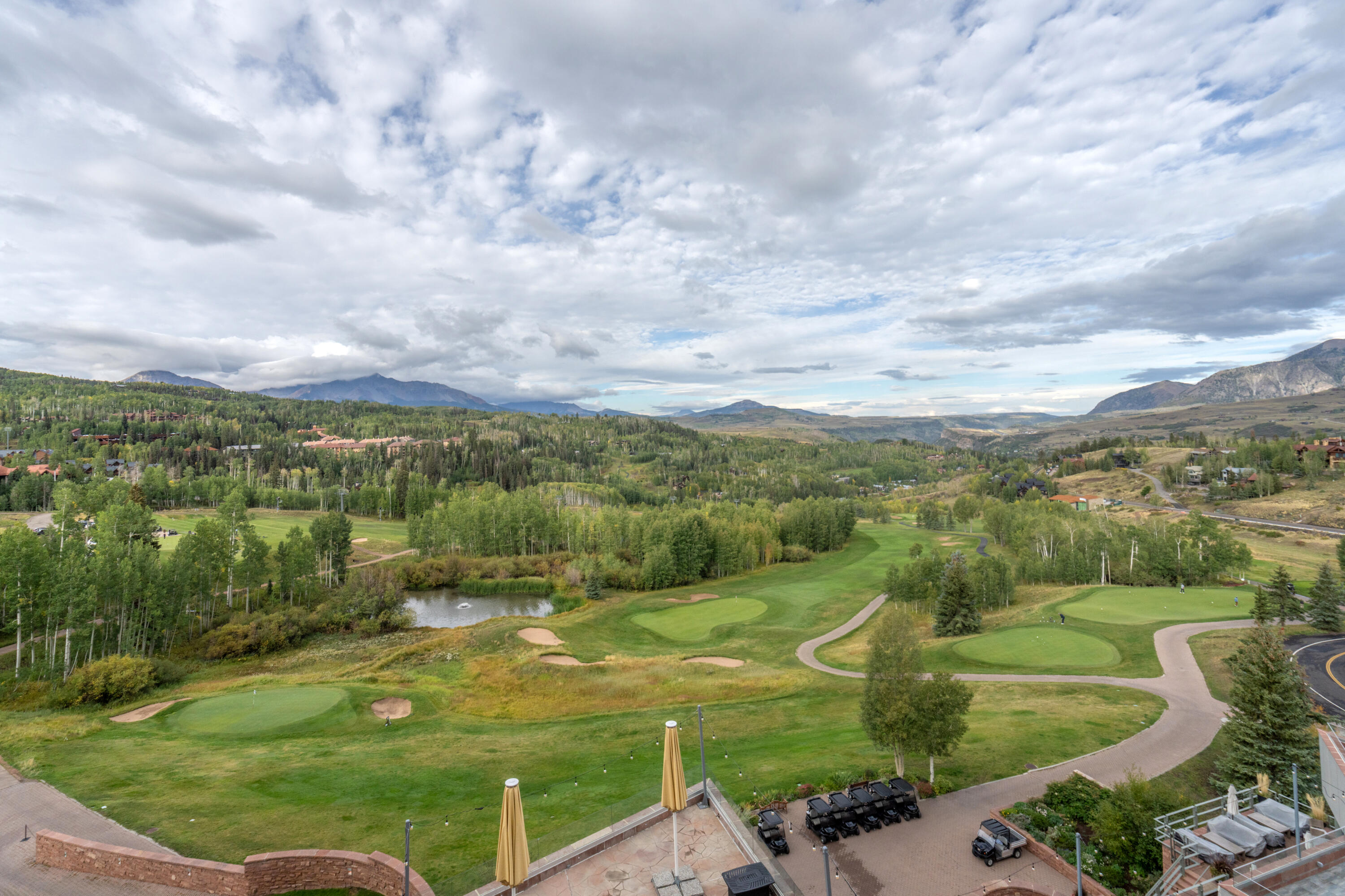 Golf course with greens and a pond, backed by forested hills and distant mountains.