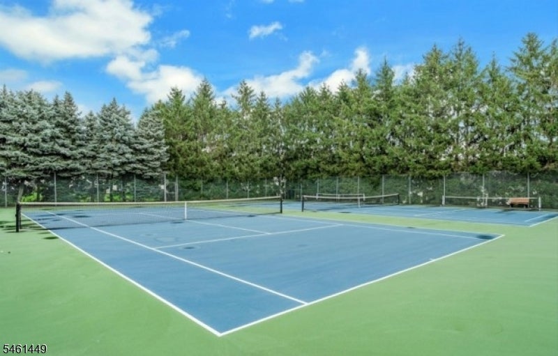 Blue tennis courts with green surrounds, chain-link fencing, and trees under a sunny sky.