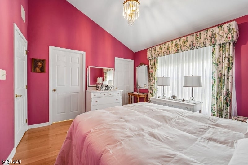 Bedroom with vivid pink walls, pale bedding, hardwood floors, and a wide window framed by floral curtains.