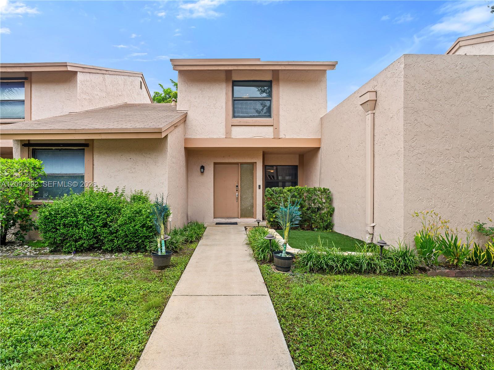Two-story beige stucco townhouse with a front walkway and landscaped lawn.