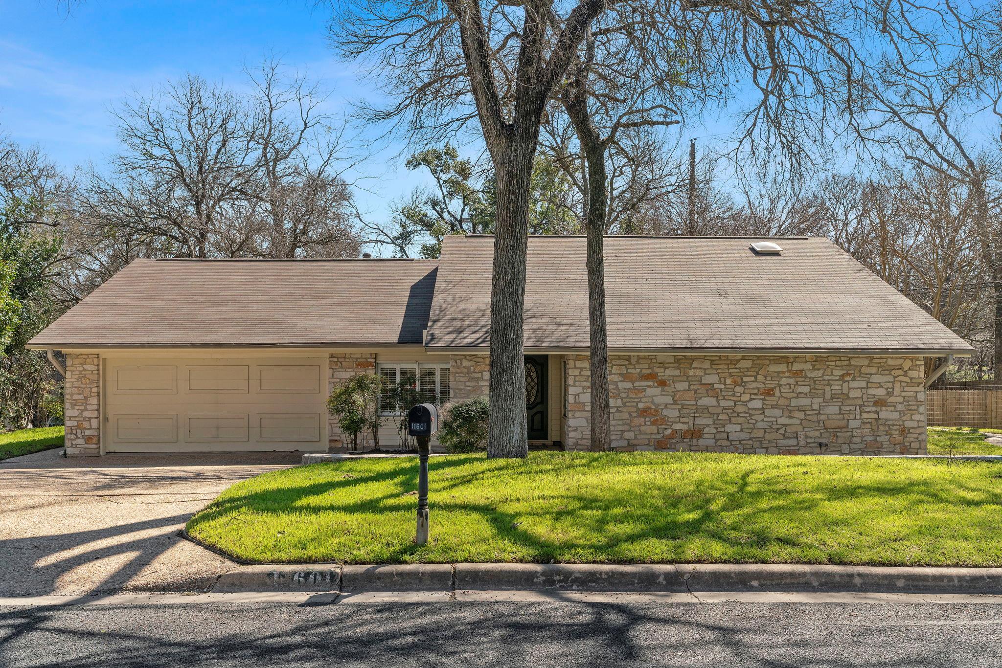 Single-story stone house with an attached garage, mature trees, and a green front lawn.
