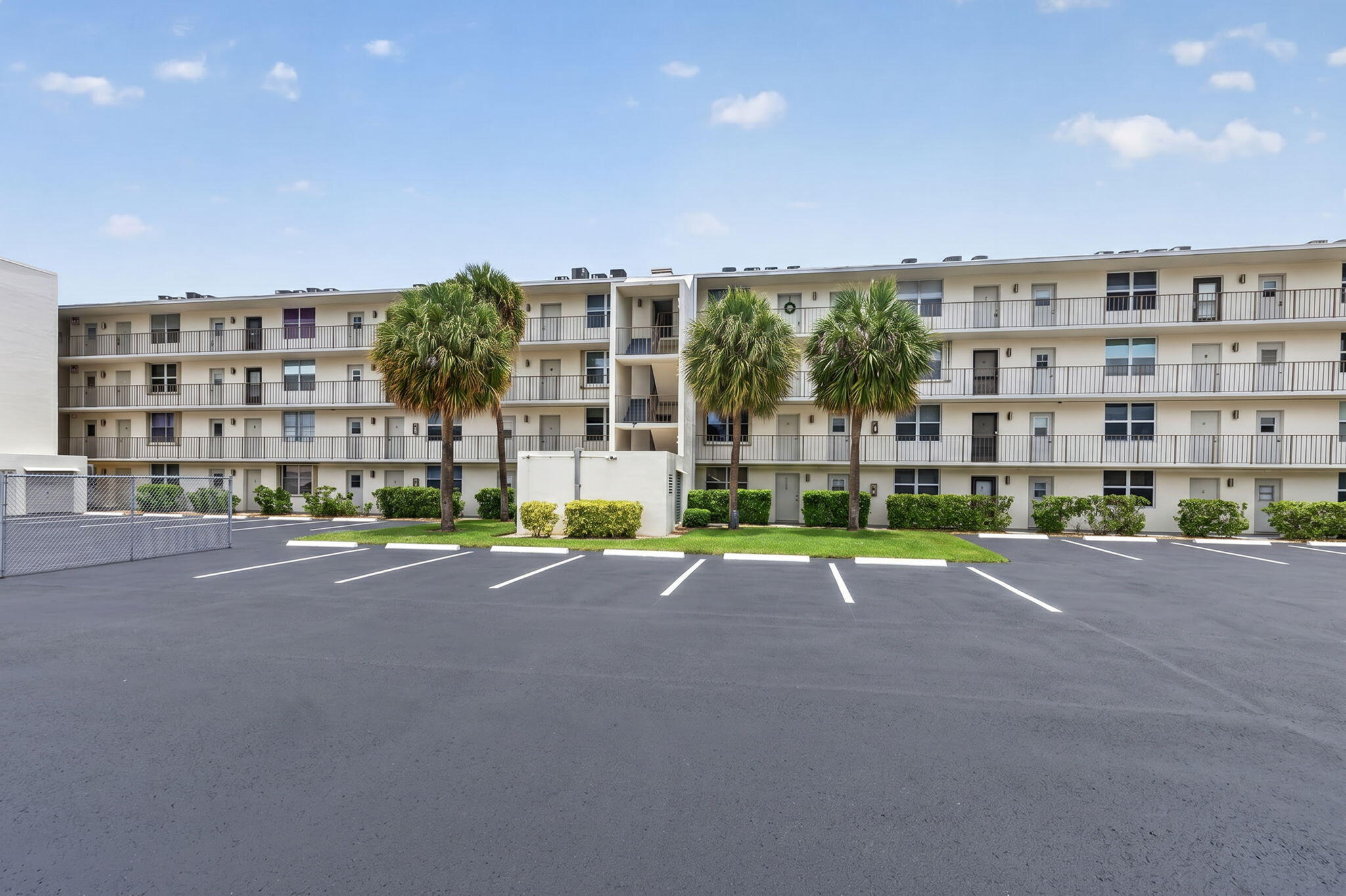 Three-story condominium building with open-air balconies and palm trees beside a paved parking lot.