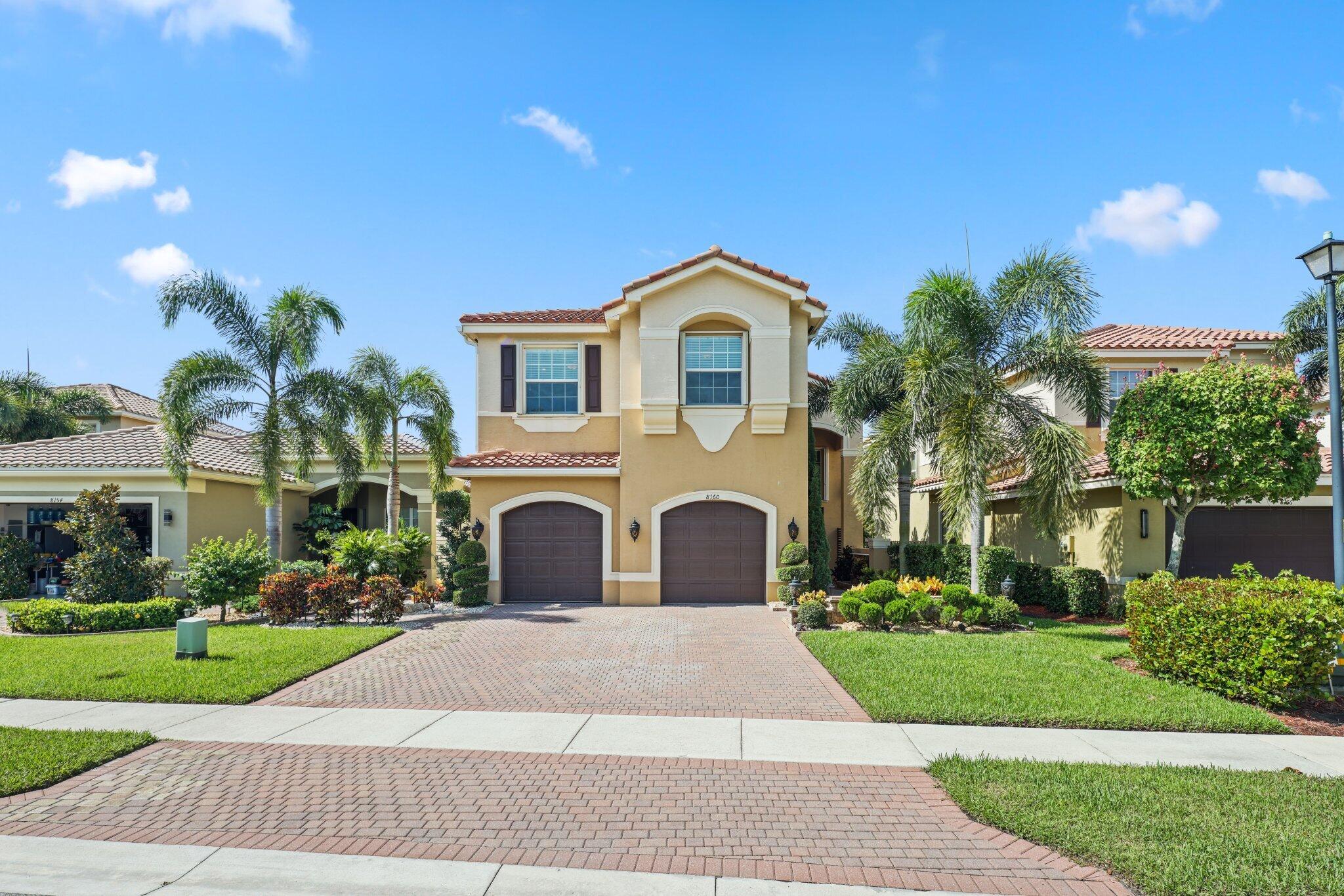 Two-story stucco home with a tile roof, twin garage doors, palm trees, and a brick-paver driveway.