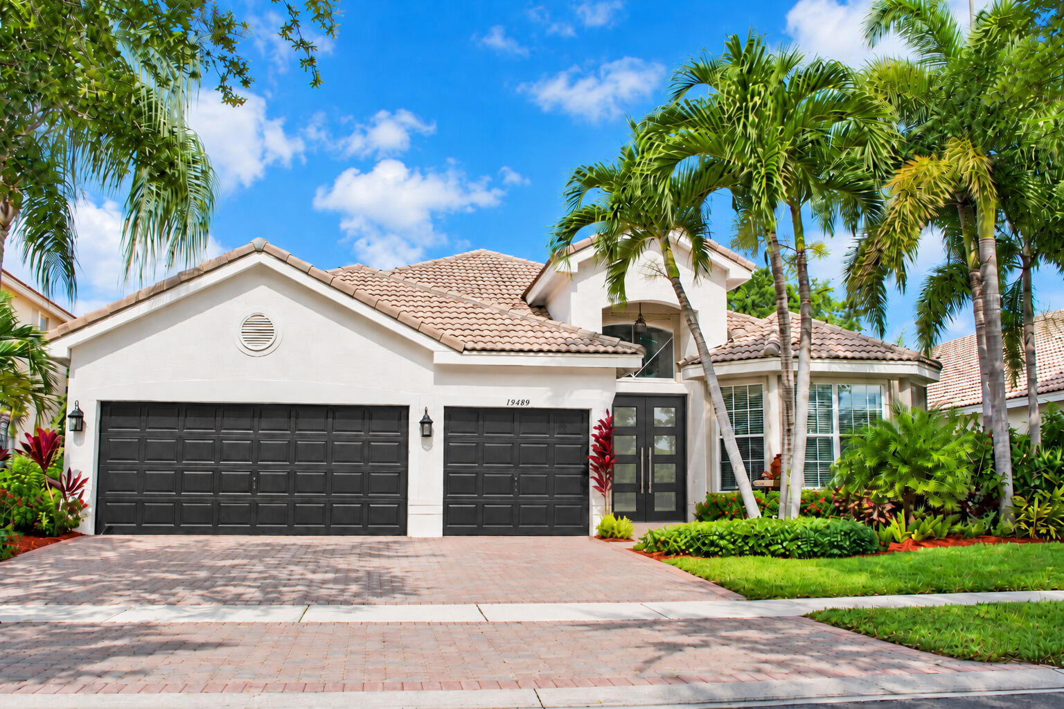 White stucco home with twin dark garage doors, terracotta roof, and palm-lined landscaping.