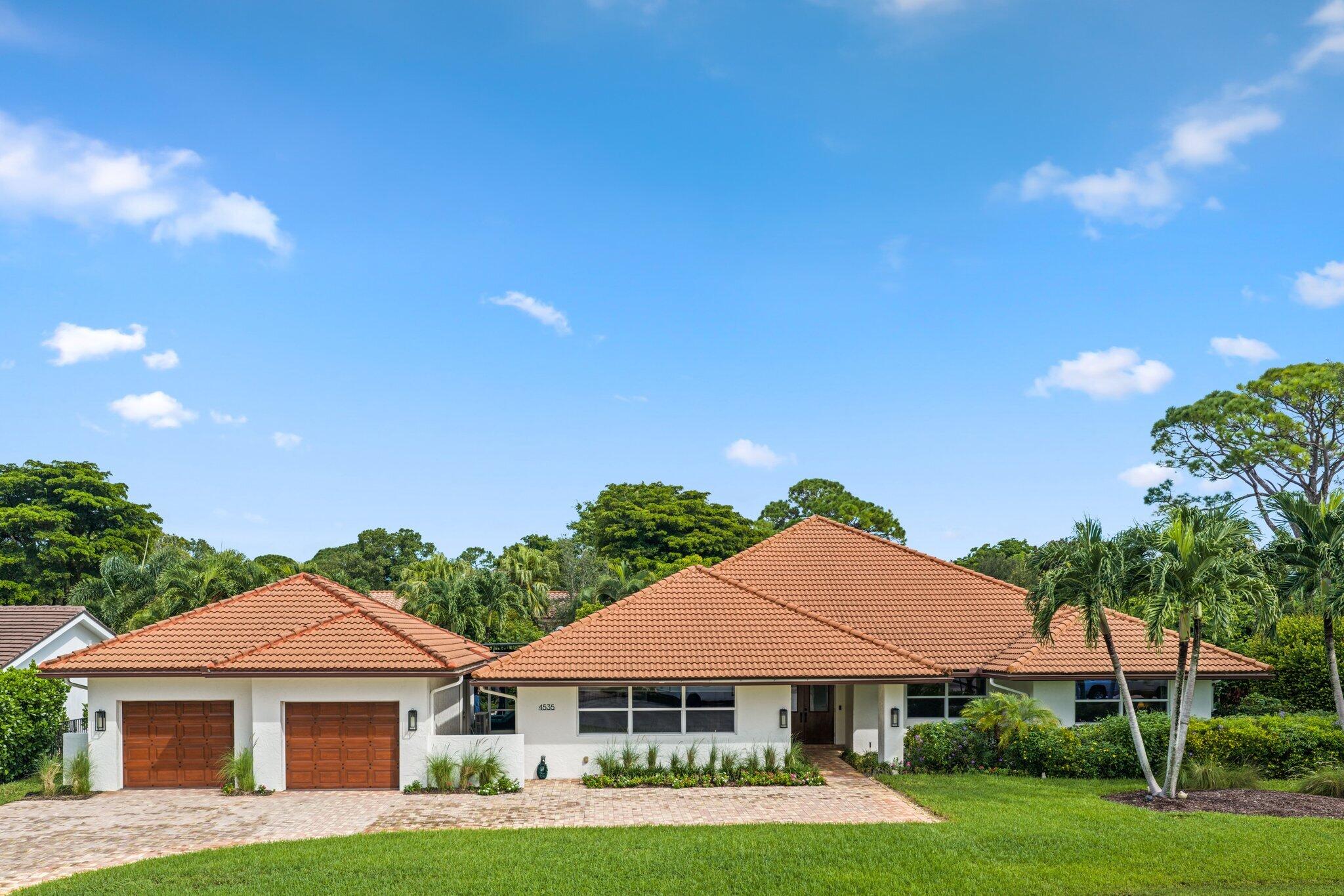 Single-story white home with a terracotta tile roof, two wooden garage doors, and tropical landscaping.