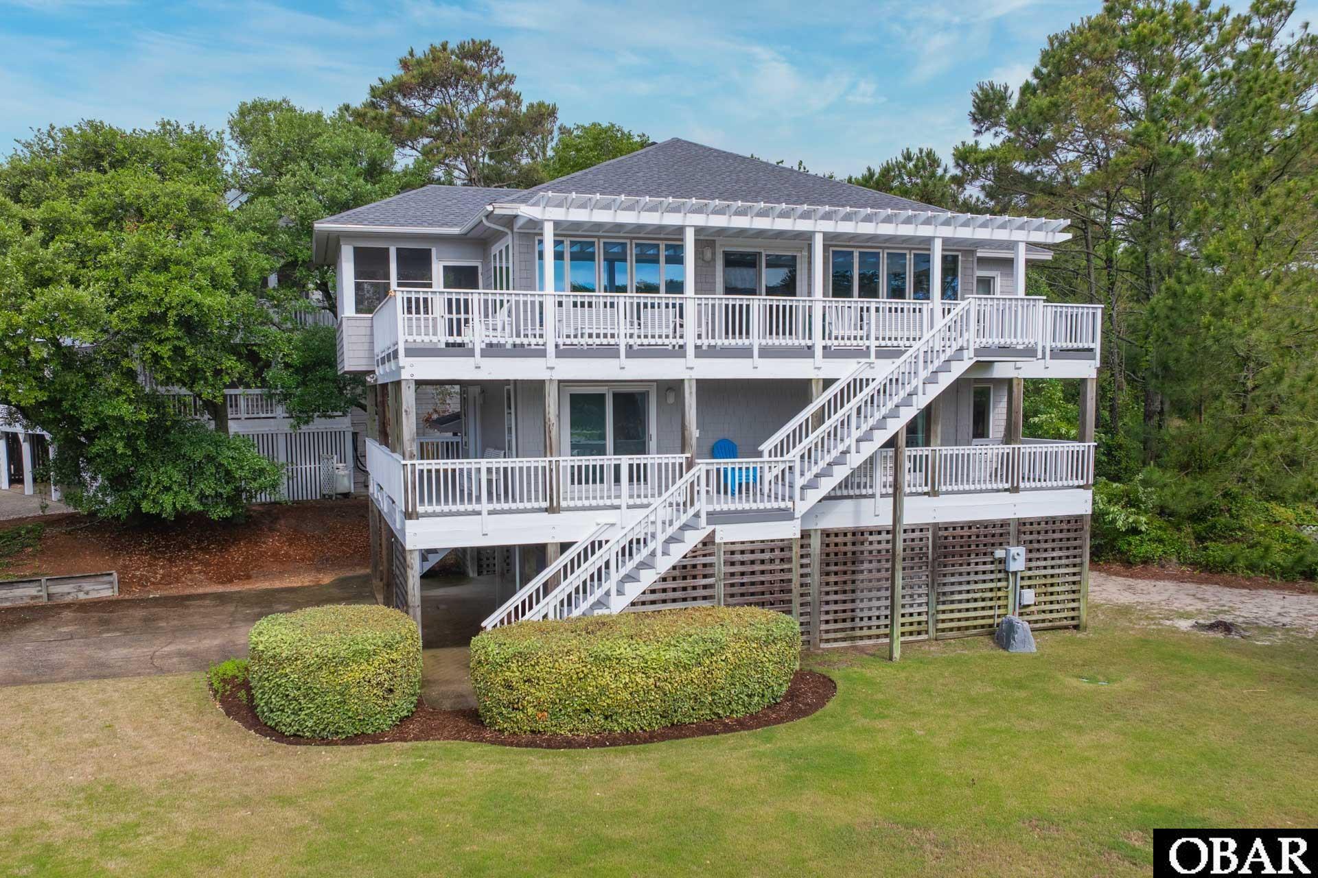 Elevated gray coastal house with white-railed decks, exterior stairs, and a pergola.