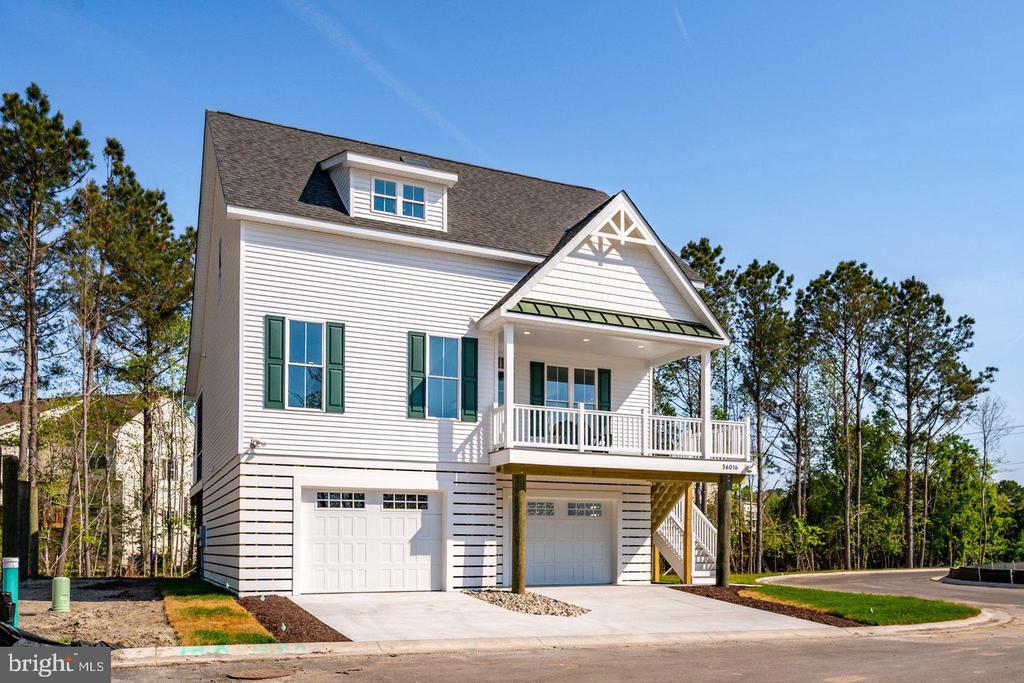 White two-story home with green shutters, a covered balcony, and two garage bays beside a wooded area.