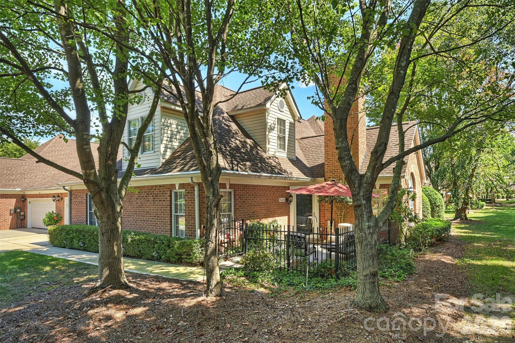 Two-story brick home shaded by mature trees, with an attached garage and fenced patio under a red umbrella.
