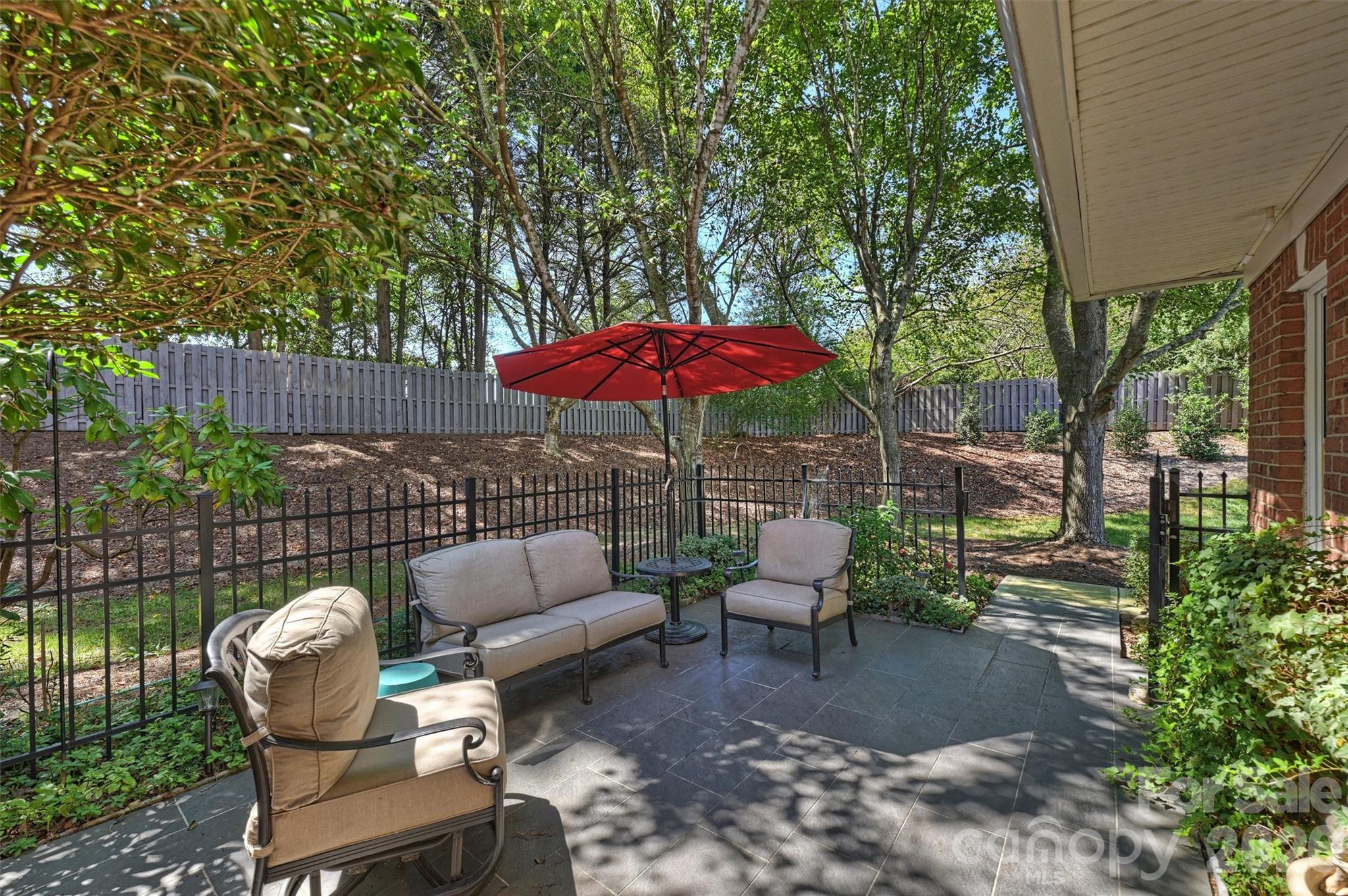 Furnished backyard patio with cushioned seating, red umbrella, and mature trees.