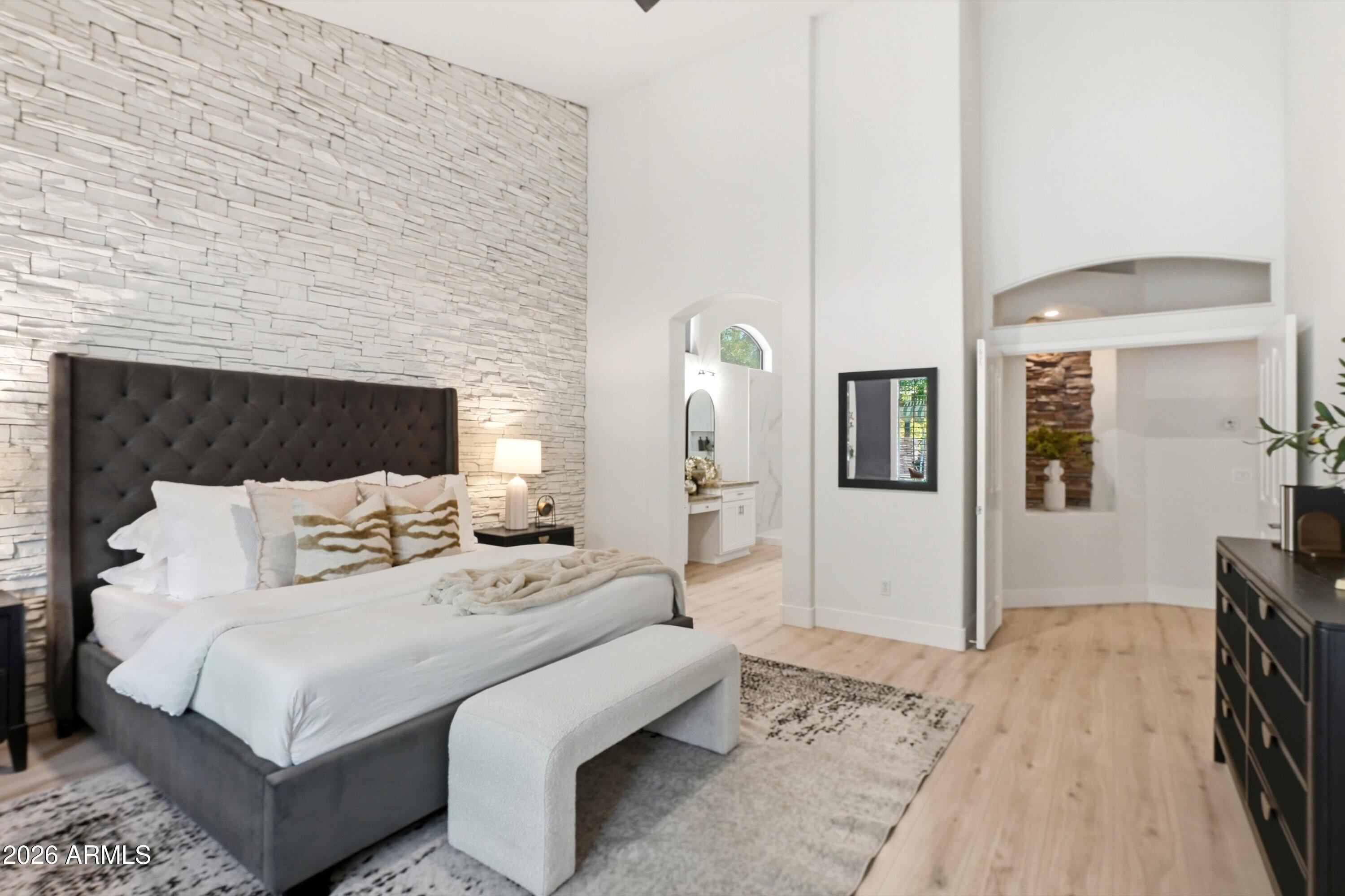 Staged primary bedroom with a dark upholstered bed, white stone accent wall, and light wood floors.