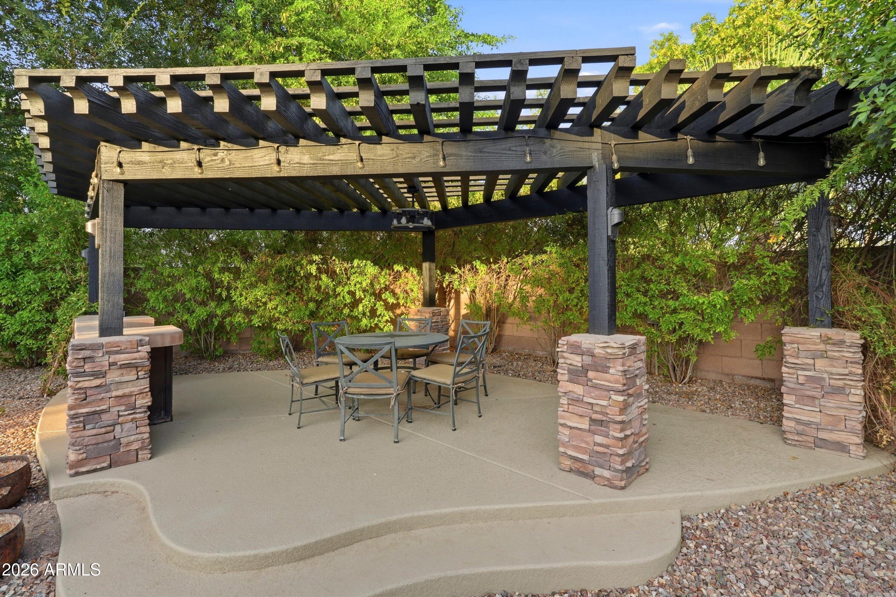 Outdoor dining patio beneath a dark wood pergola with string lights and stone-clad column bases.