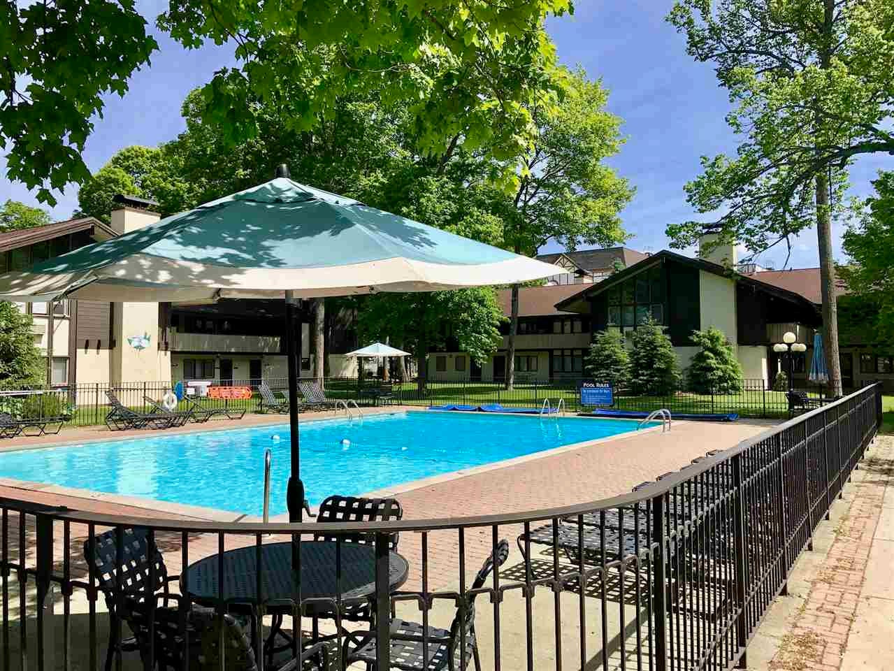 Condominium pool with a brick deck, shade umbrellas, lounge chairs, and leafy trees.