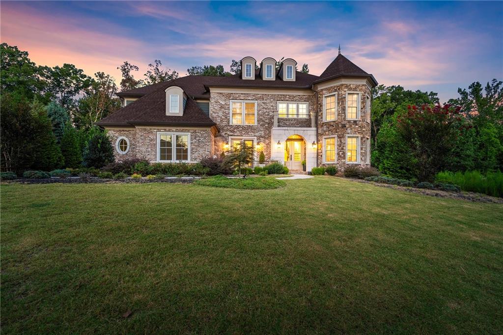 Two-story brick-and-stone house with a turret, glowing windows, and broad front lawn at dusk.
