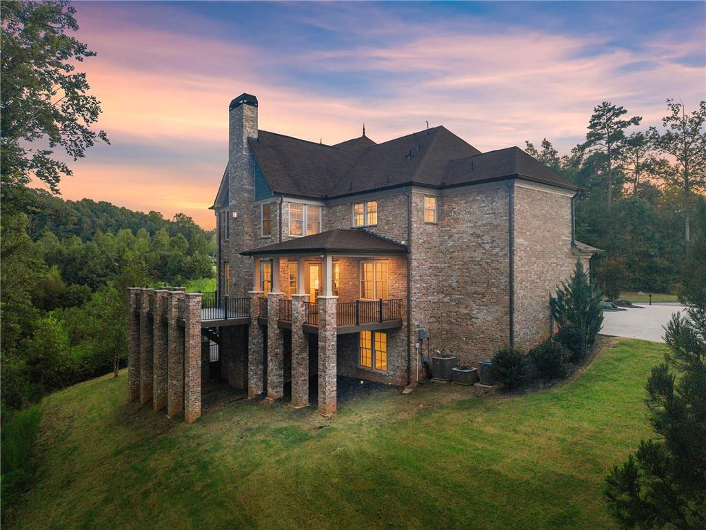 Brick house with a raised deck and stone chimney, surrounded by trees at dusk.