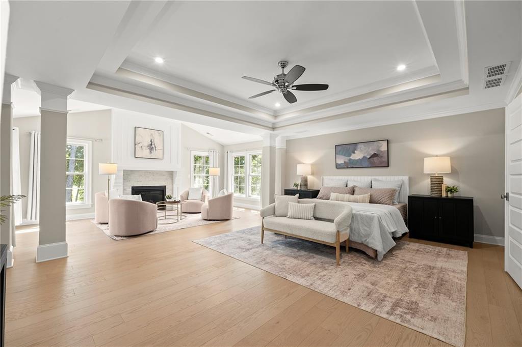 Staged bedroom with a tray ceiling, fireplace, and adjoining sitting area with tall windows.