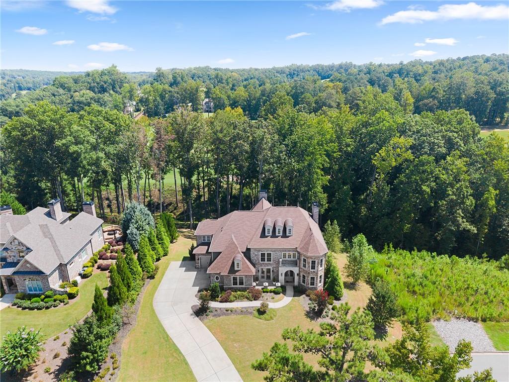 Aerial view of a large stone home, curved driveway, landscaped grounds, and surrounding woodland.
