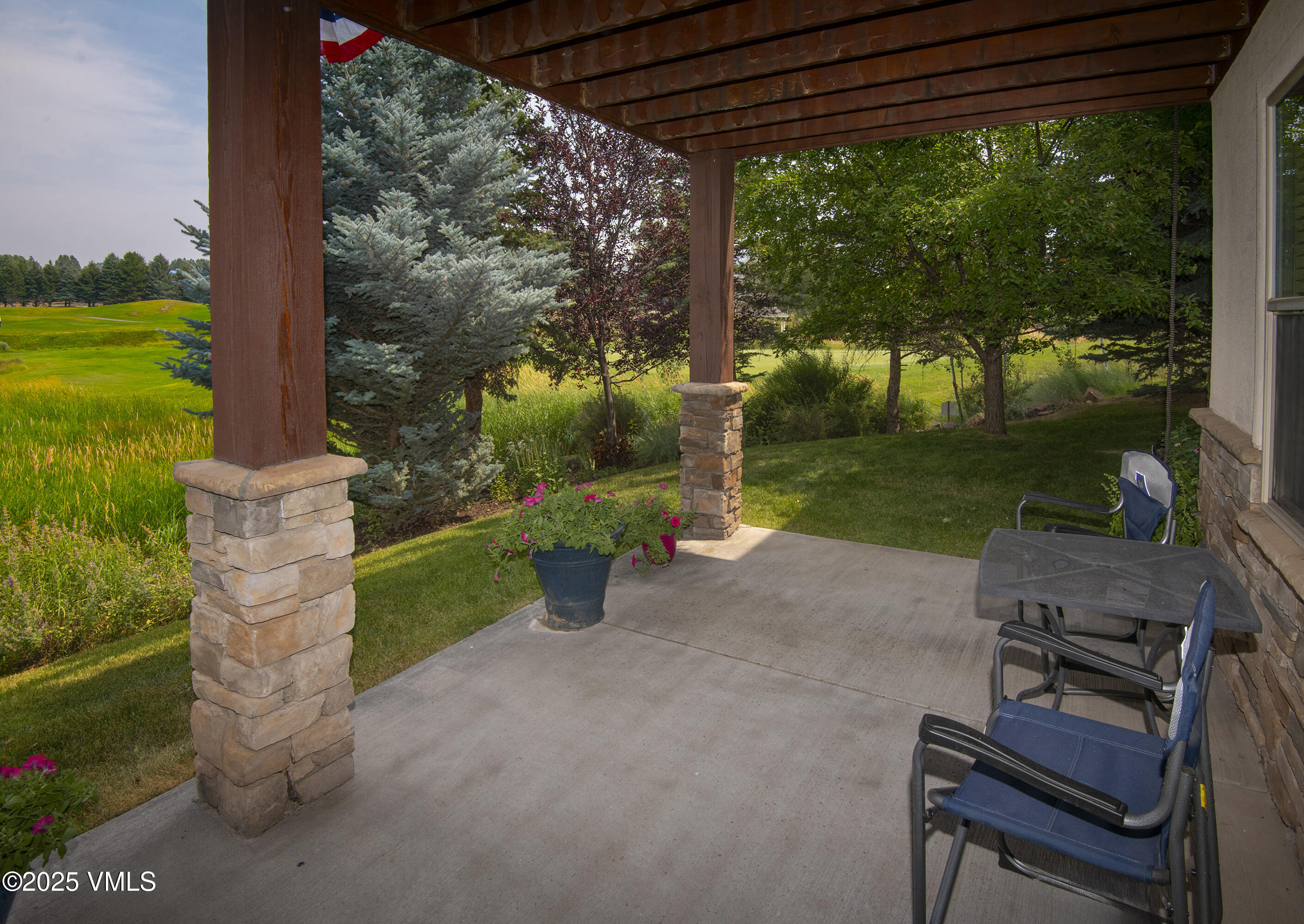 Covered patio with blue chairs and flowering planters overlooking a landscaped lawn.