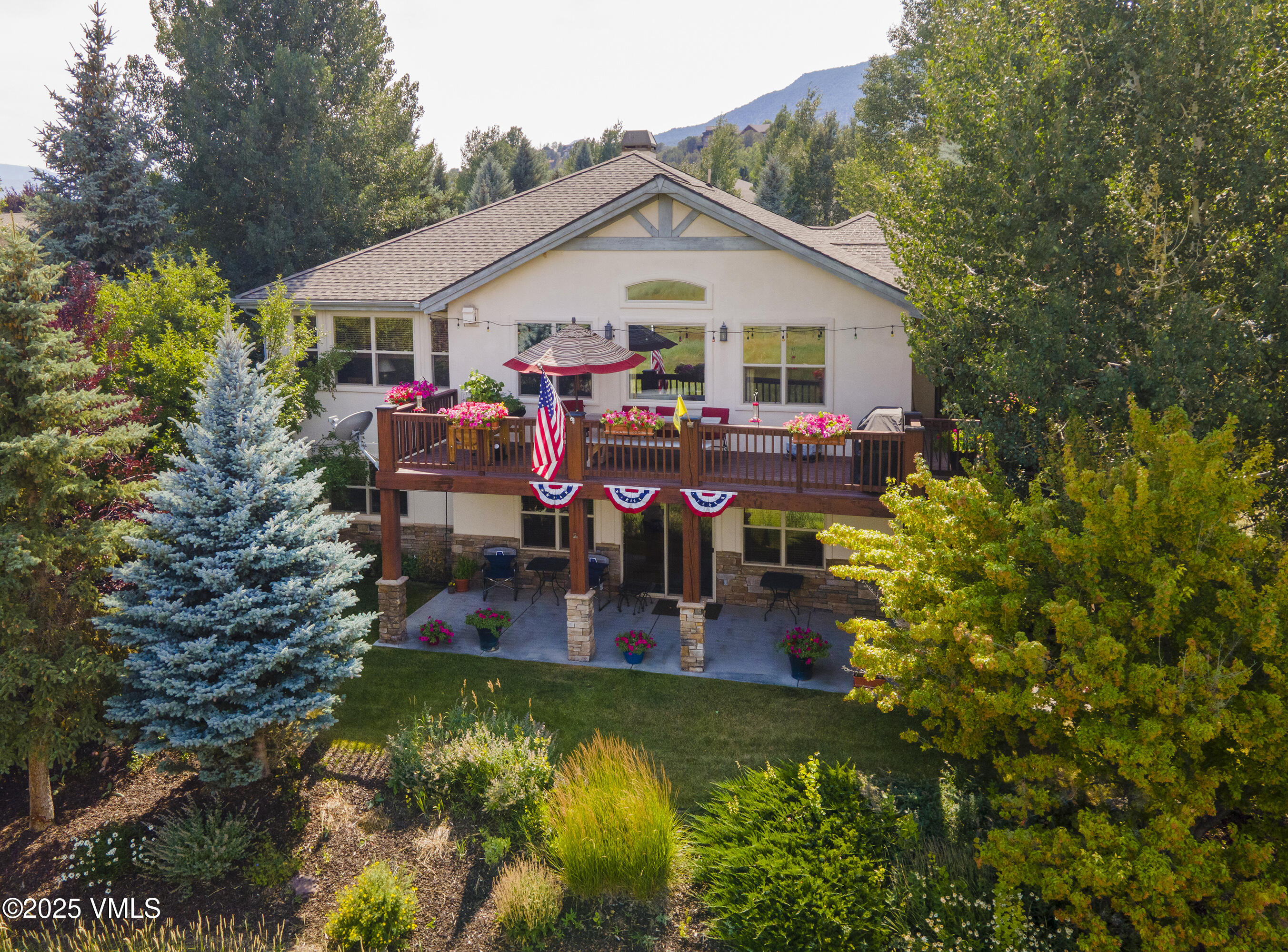 Two-story home with a flower-lined deck and covered patio, surrounded by mature trees and landscaped gardens.
