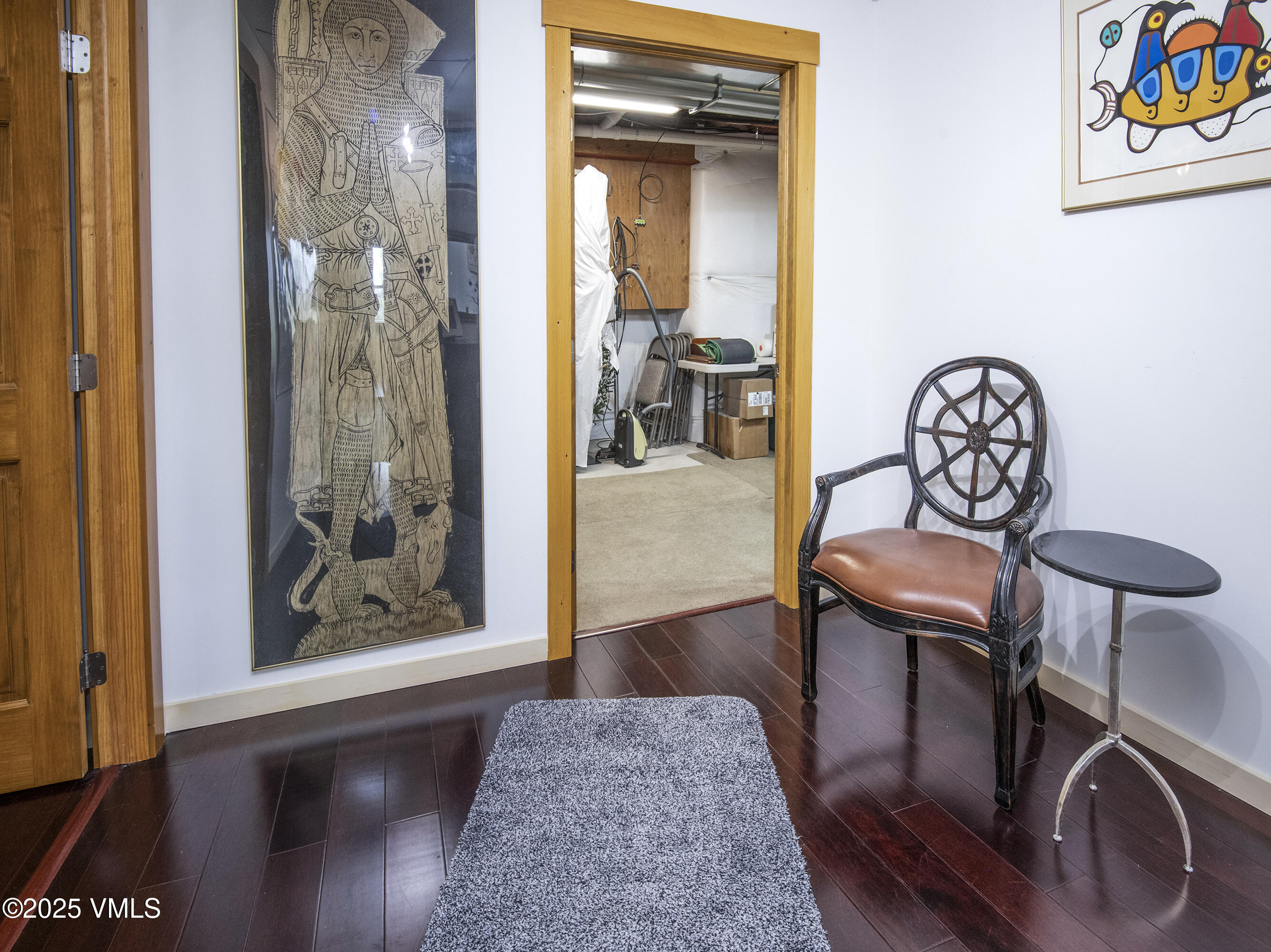 Entryway with dark hardwood floors, framed artwork, and a chair beside an open storage-room doorway.