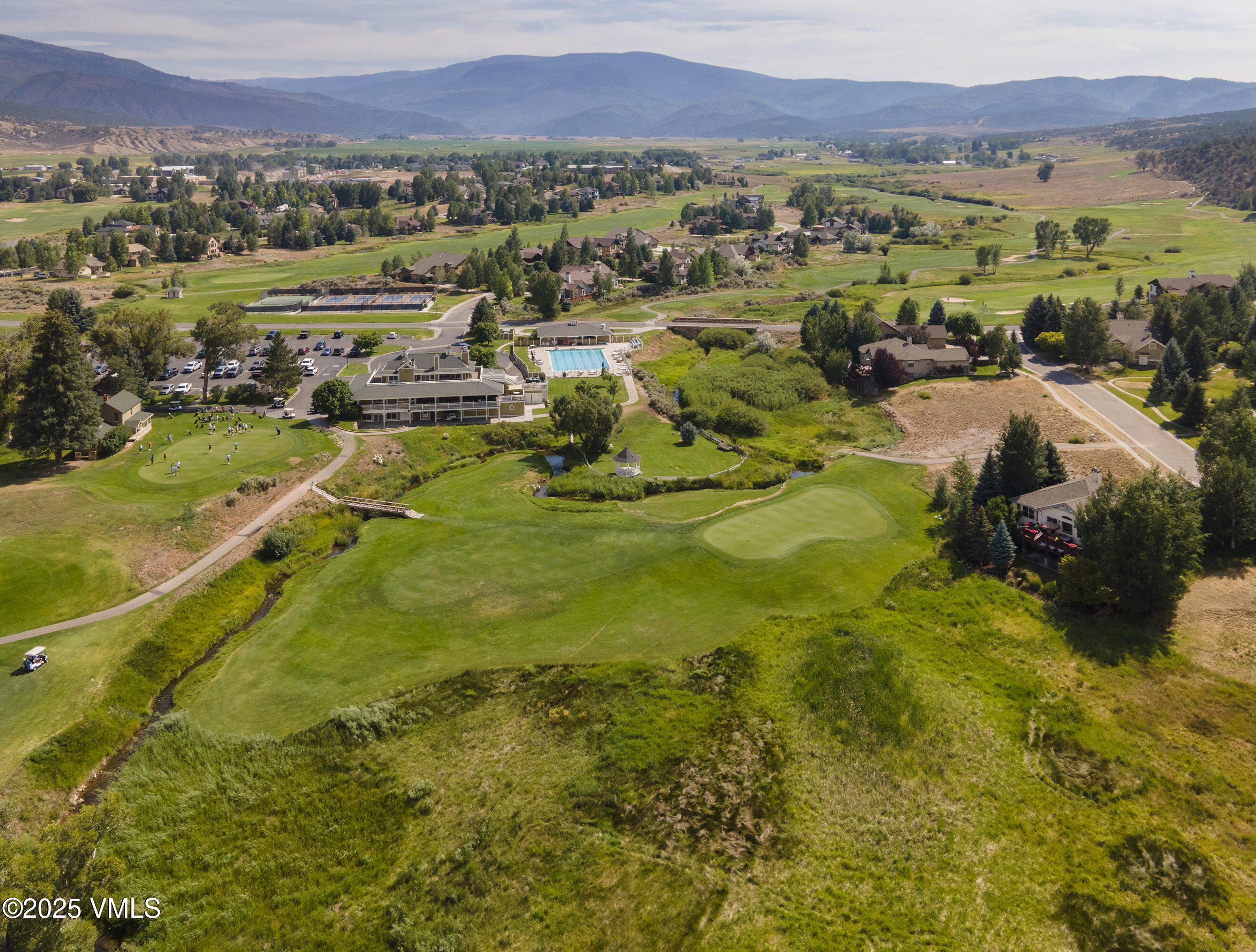 Aerial view of a golf course community with a clubhouse, swimming pool, creek, and mountain backdrop.