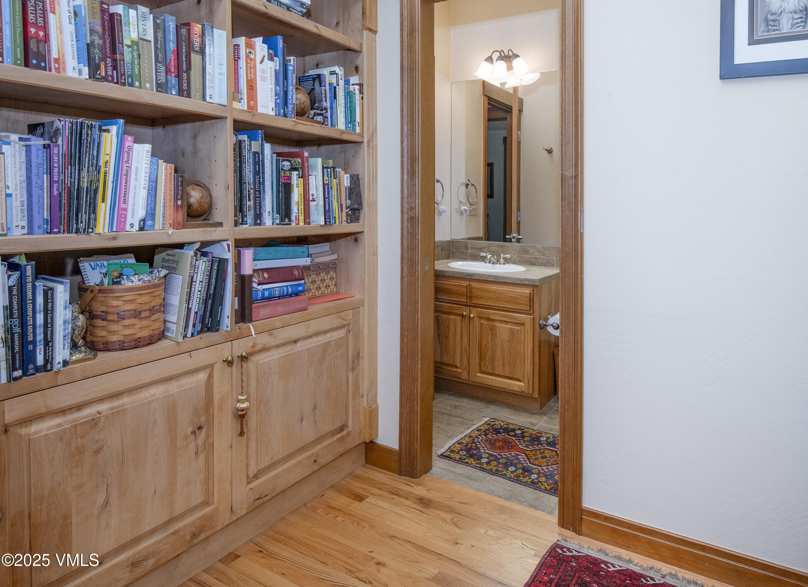 Built-in wood bookcase beside an open bathroom doorway with a wood vanity and patterned rugs.