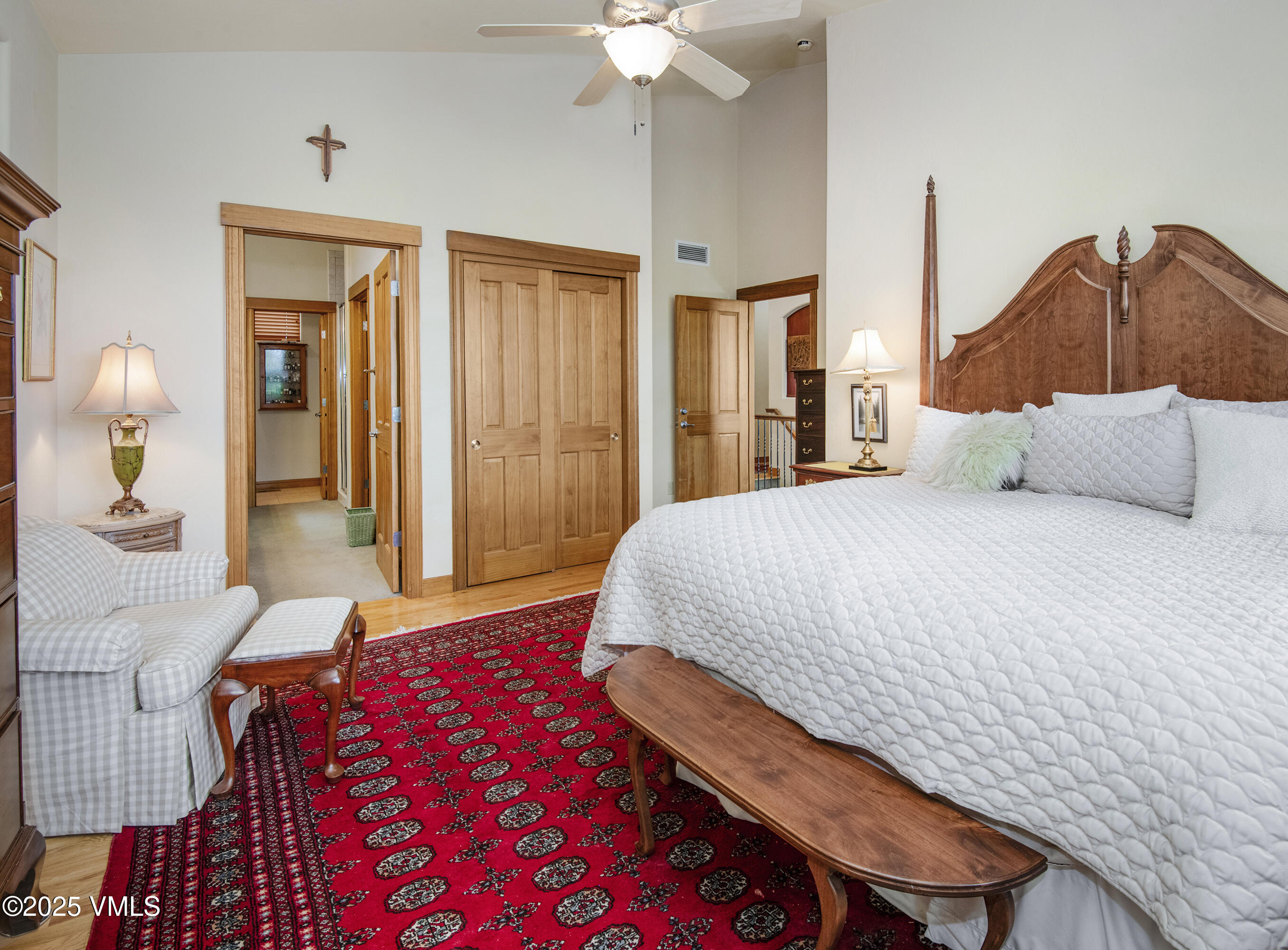 Bedroom with an ornate carved wood headboard, white quilt, red patterned rug, and checkered armchair.