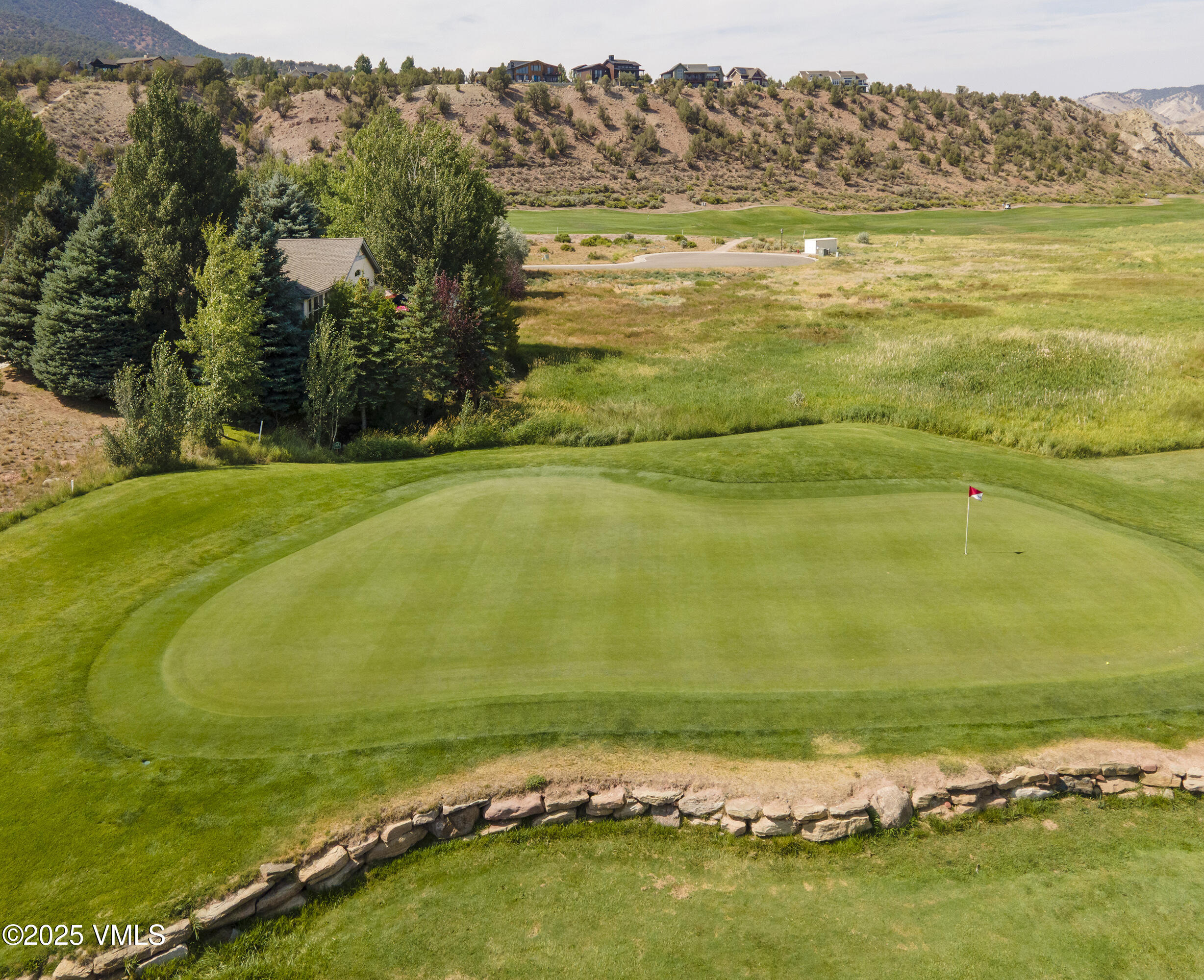 Golf course green with a red flag and curved stone wall, backed by homes and dry hills.