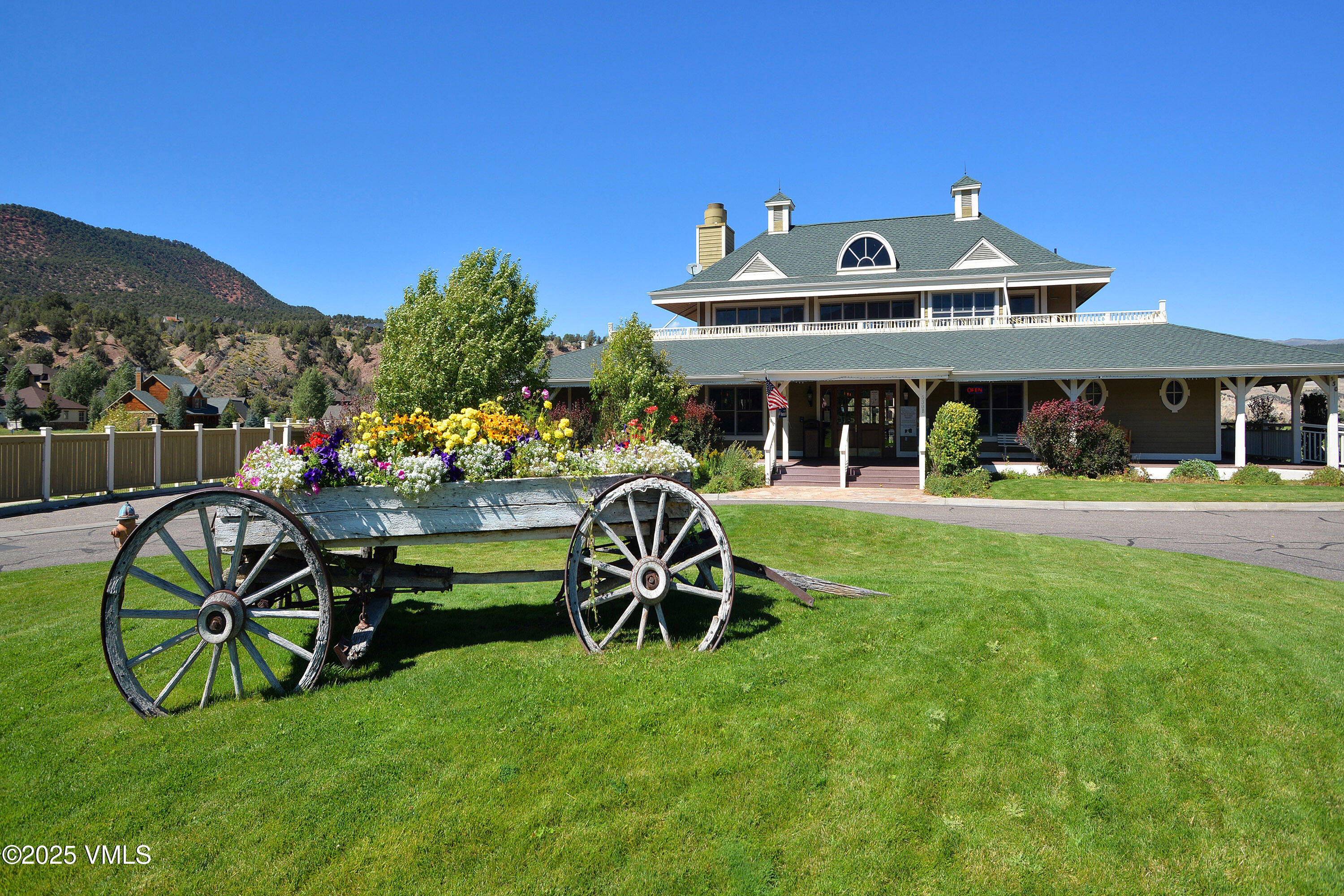 Large green-roofed building with a covered porch and flower-filled wagon planter, backed by a wooded hillside.