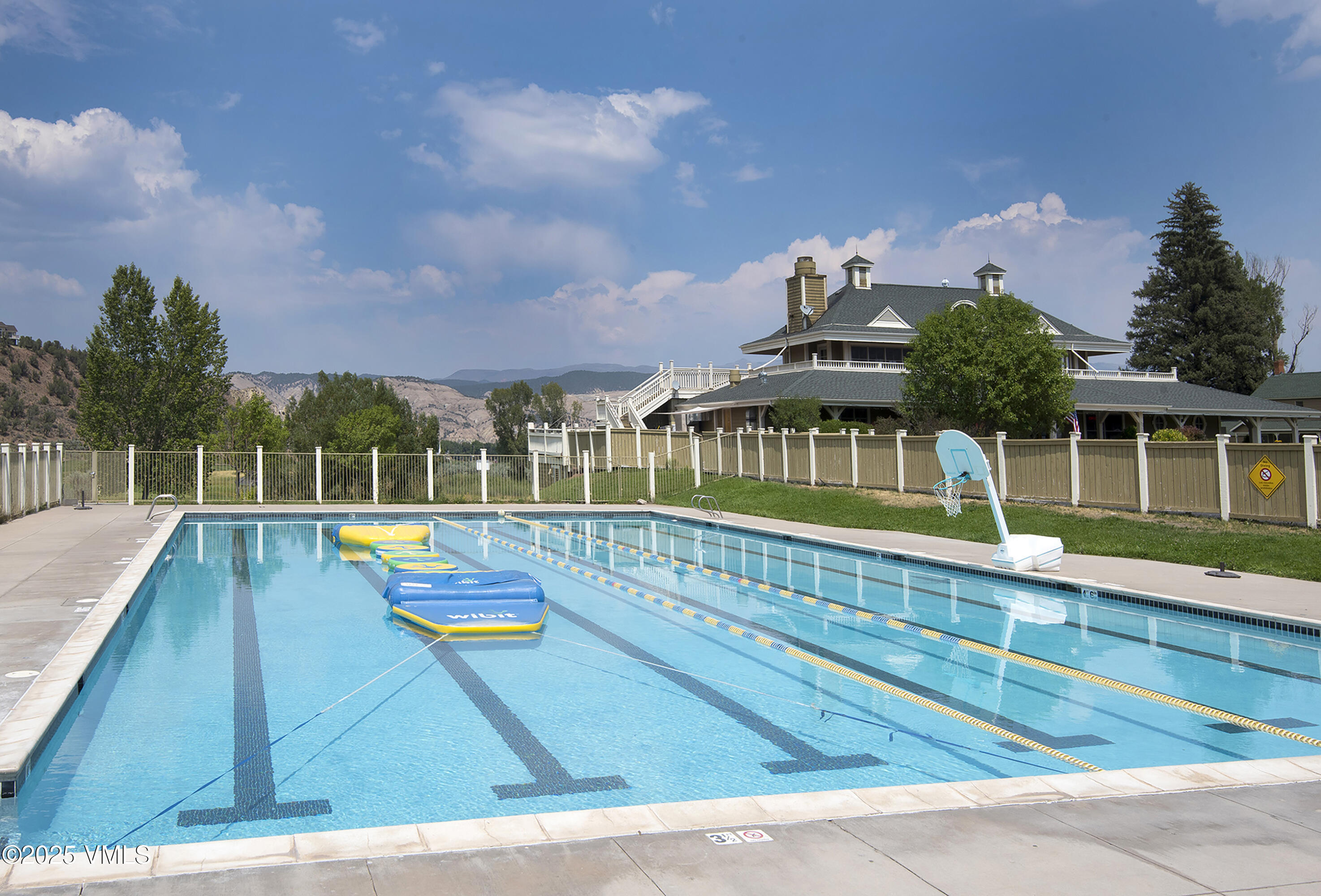 Outdoor pool with lane markings and floating mats, backed by a fenced recreation area and mountain views.