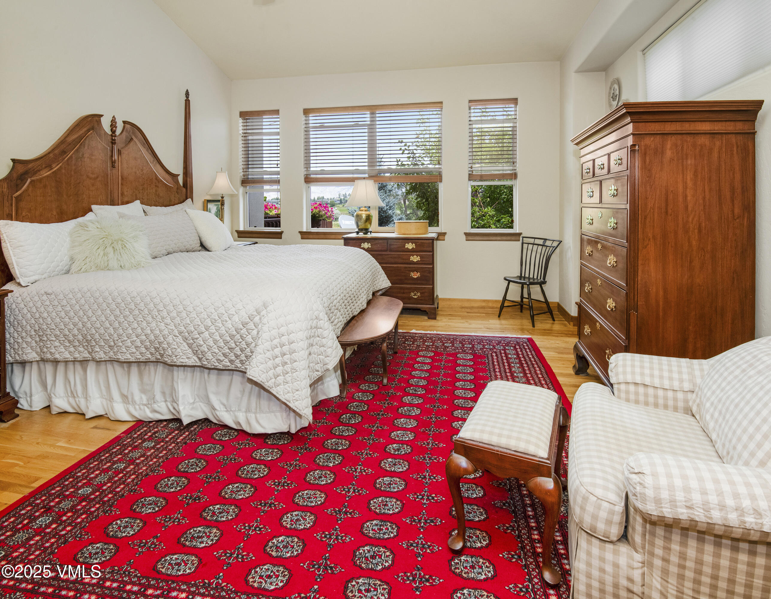 Bedroom with carved wooden bed, red patterned rug, and windows overlooking greenery.